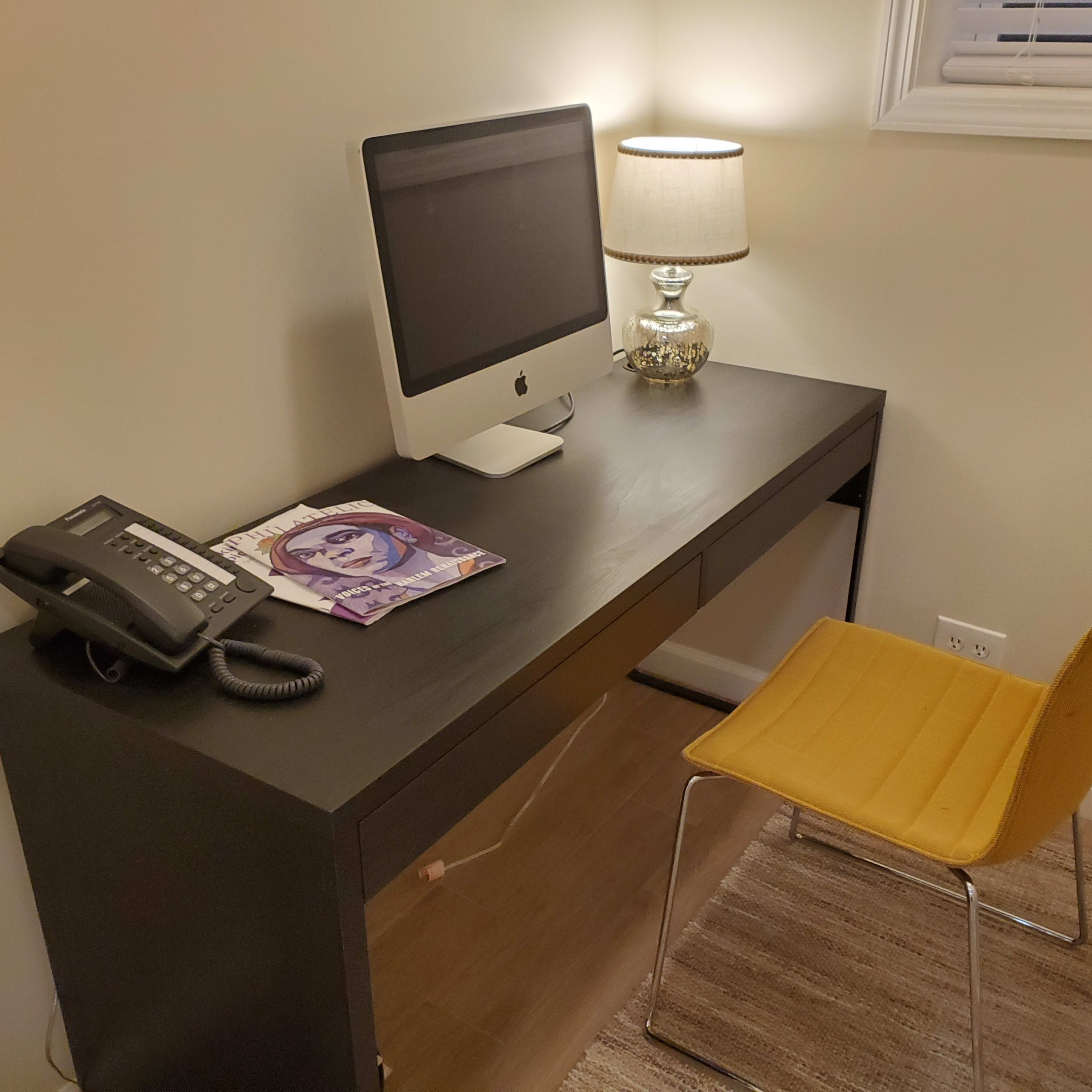 A black desk with a computer, a phone, a magazine, a yellow chair, and a lamp is positioned in a well-lit room.