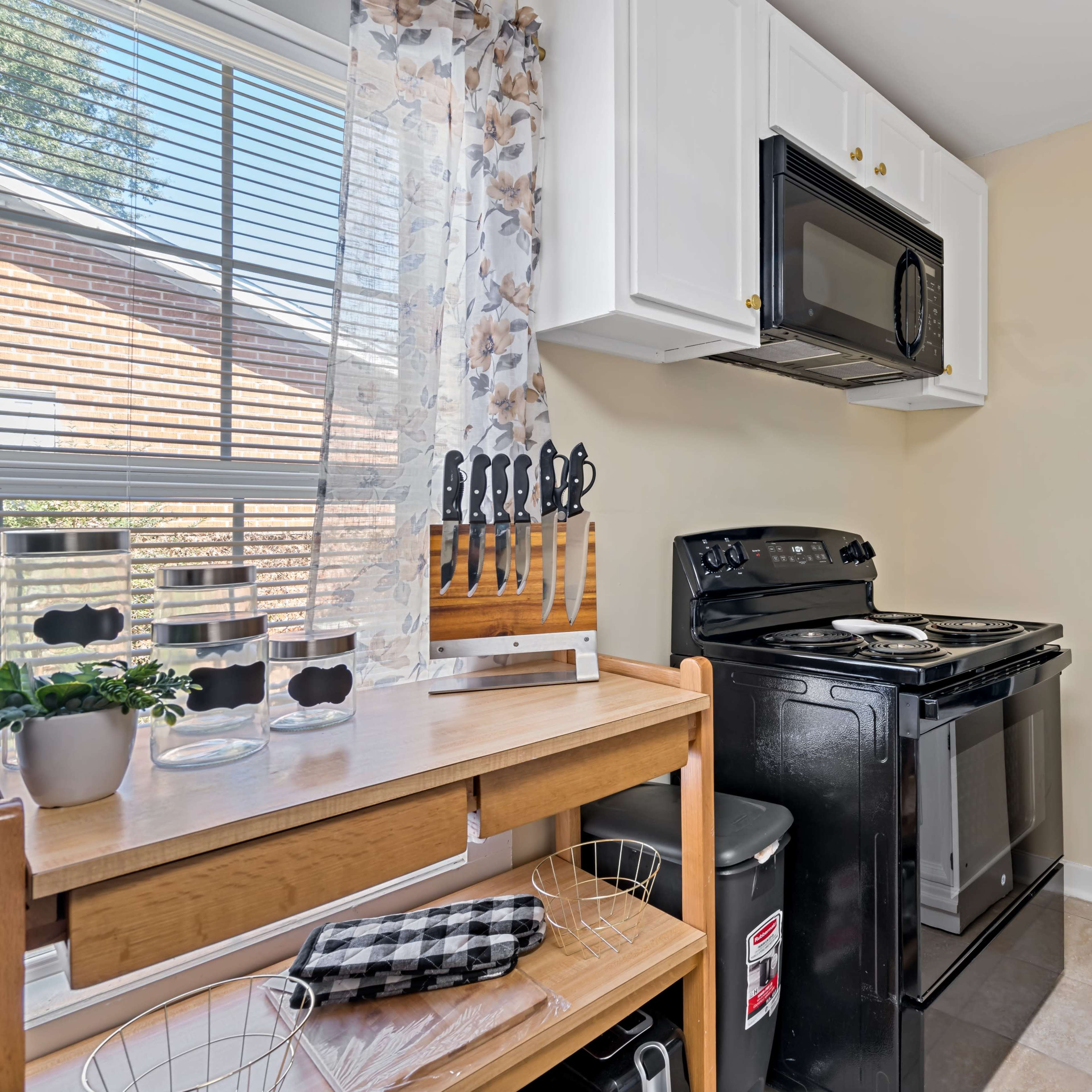 A small kitchen space featuring a black cooking range, a microwave mounted above it, a shelving unit with kitchen supplies, and a wooden table with knives and a plant.