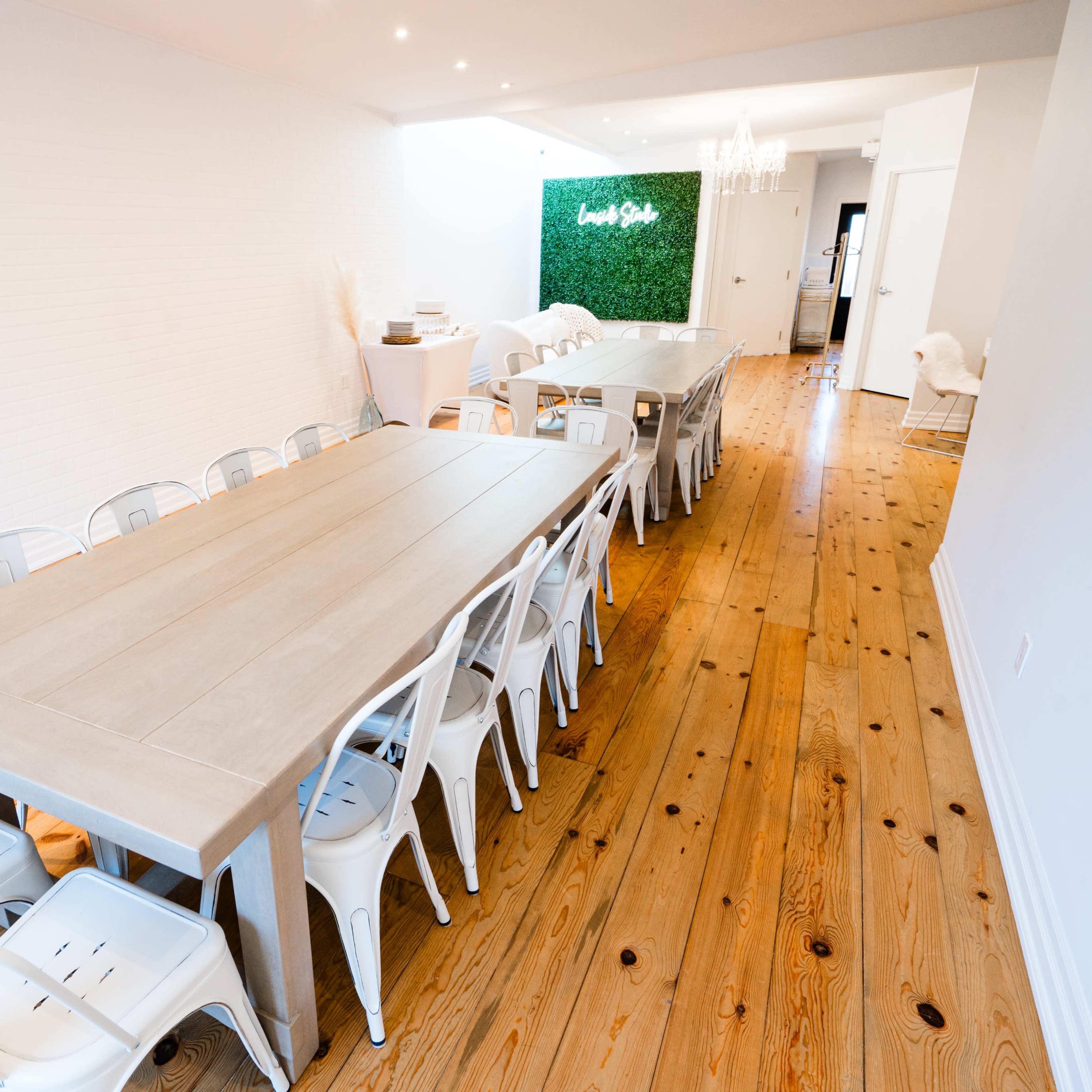 A long rectangular table with white chairs is set up in a spacious room with wooden floors and green wall decor at one end.