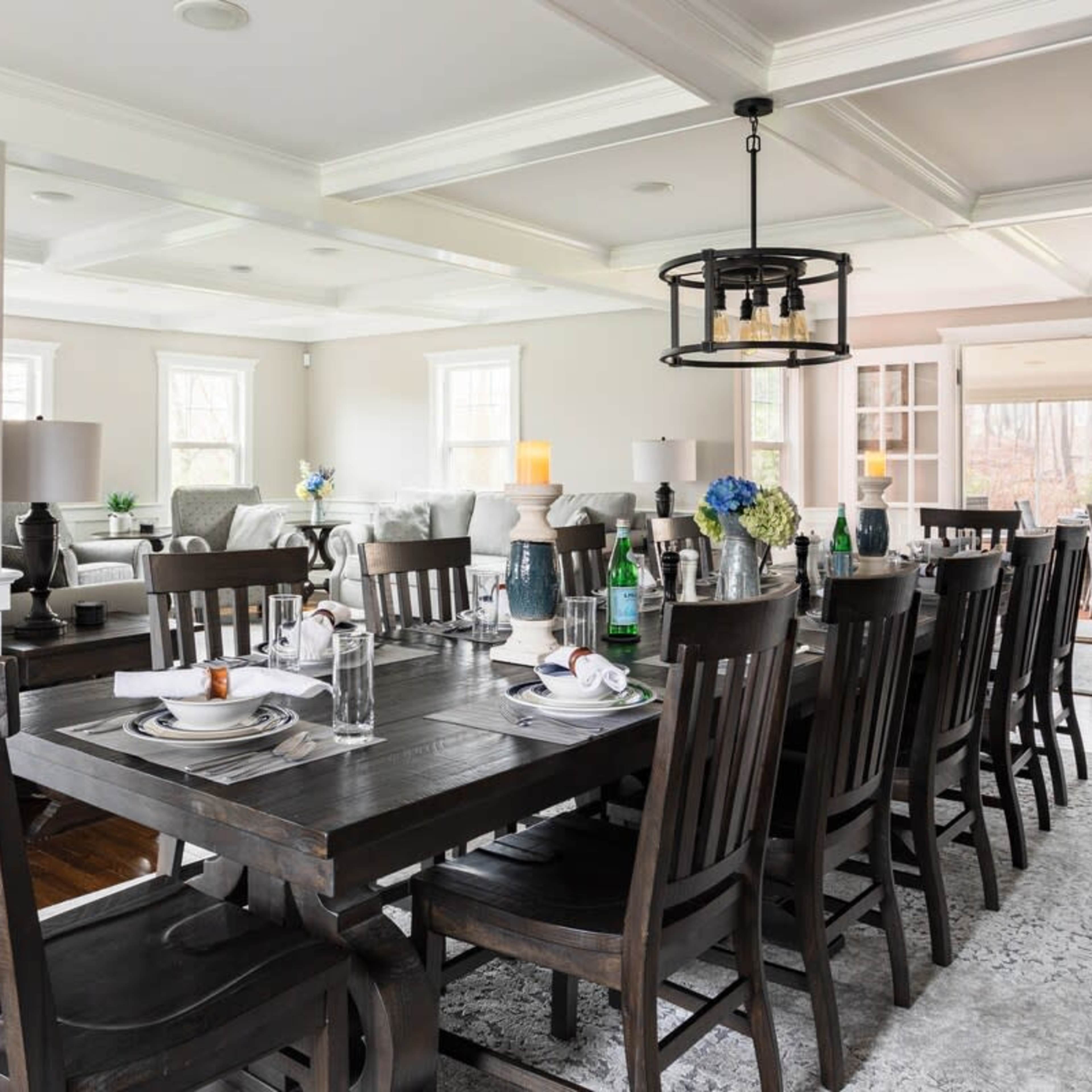 A long wooden dining table is set for a meal, surrounded by dark chairs, with a view into a bright living area through open doors.