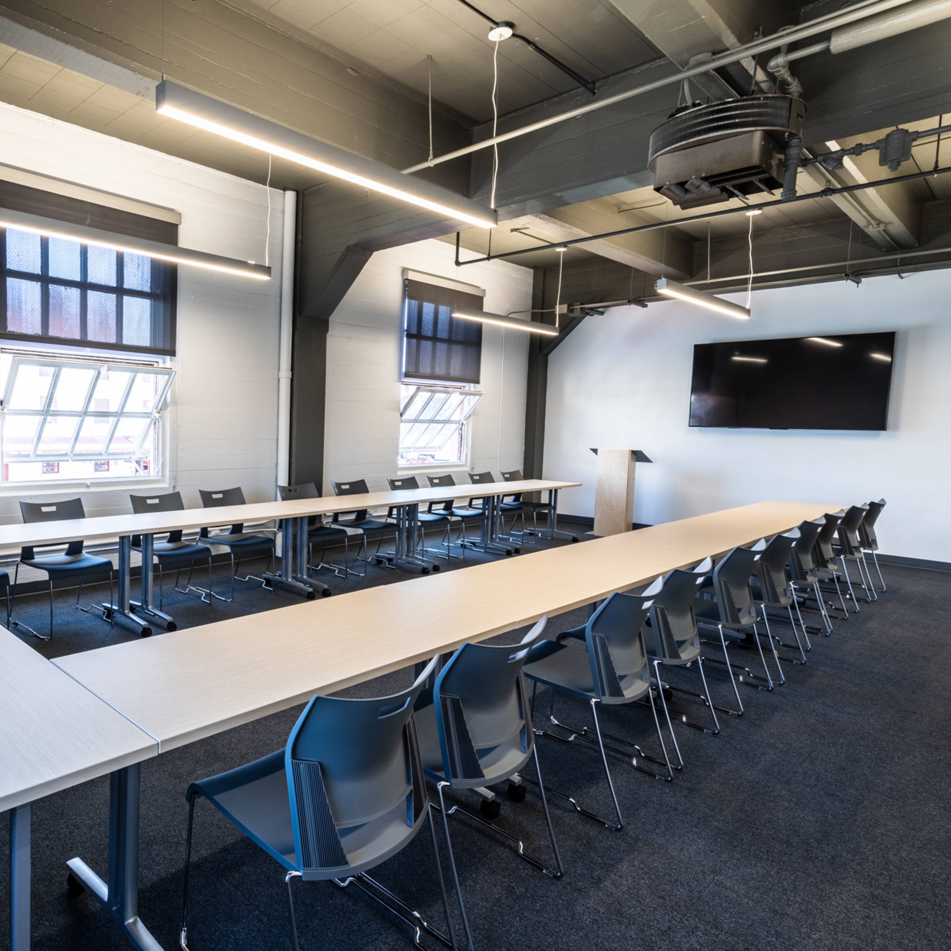A modern conference room features a long table arranged in a U-shape with gray chairs and large windows.