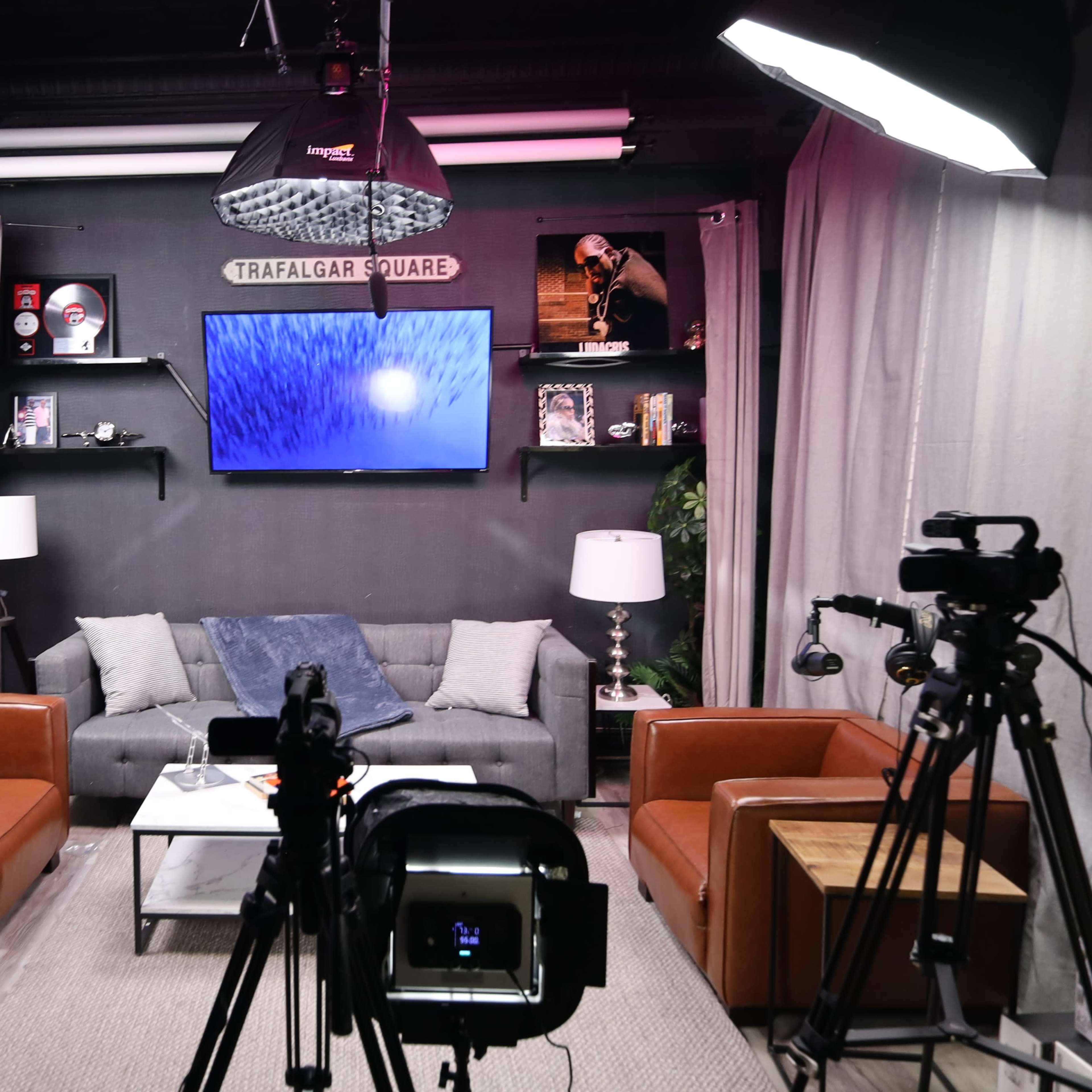 A modern studio setup with two cameras aimed at a seating area featuring a gray couch, two brown chairs, and a screen displaying visuals on the wall.