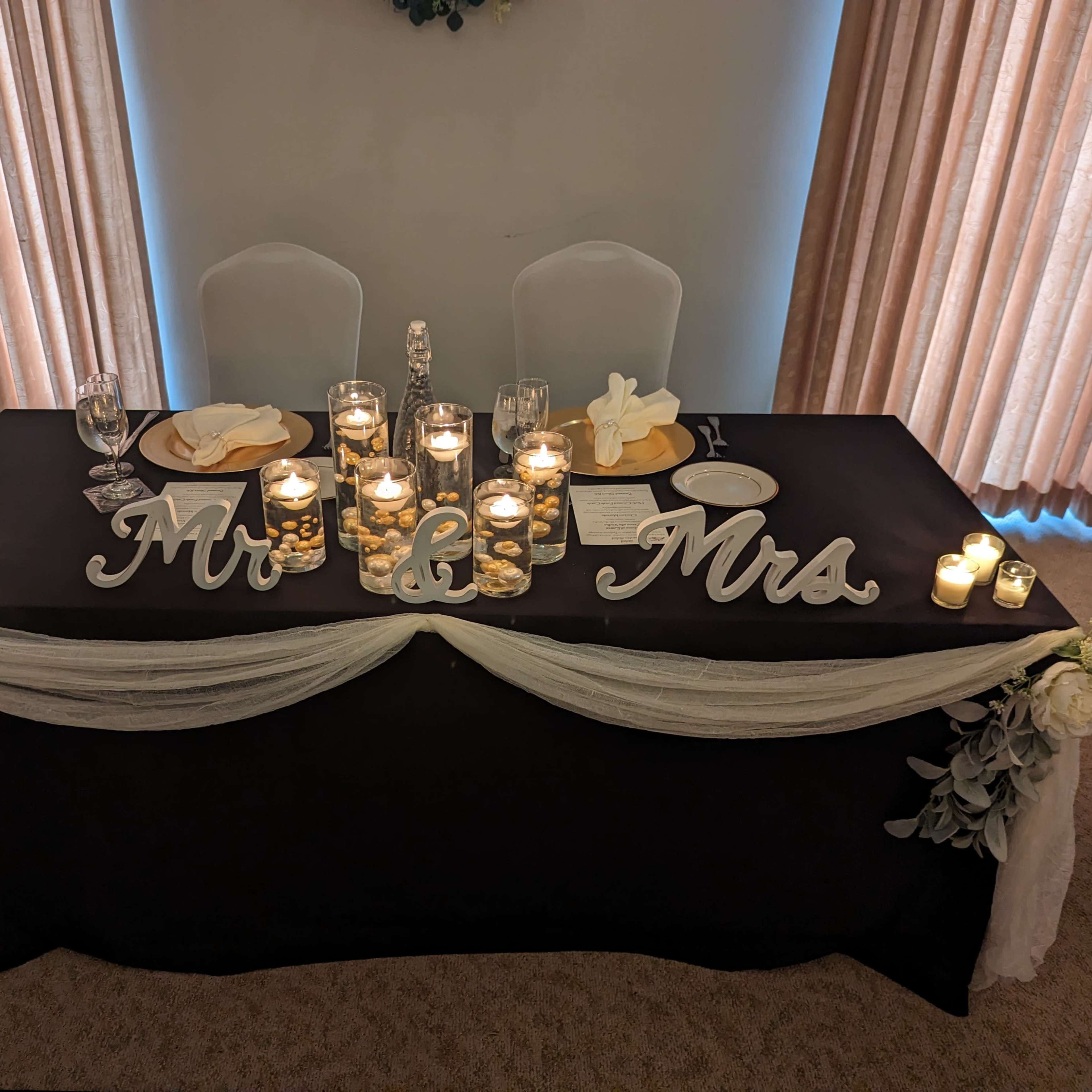 A table set for a wedding reception features a black tablecloth, candles, floral arrangements, and decorative "Mr & Mrs" signs.