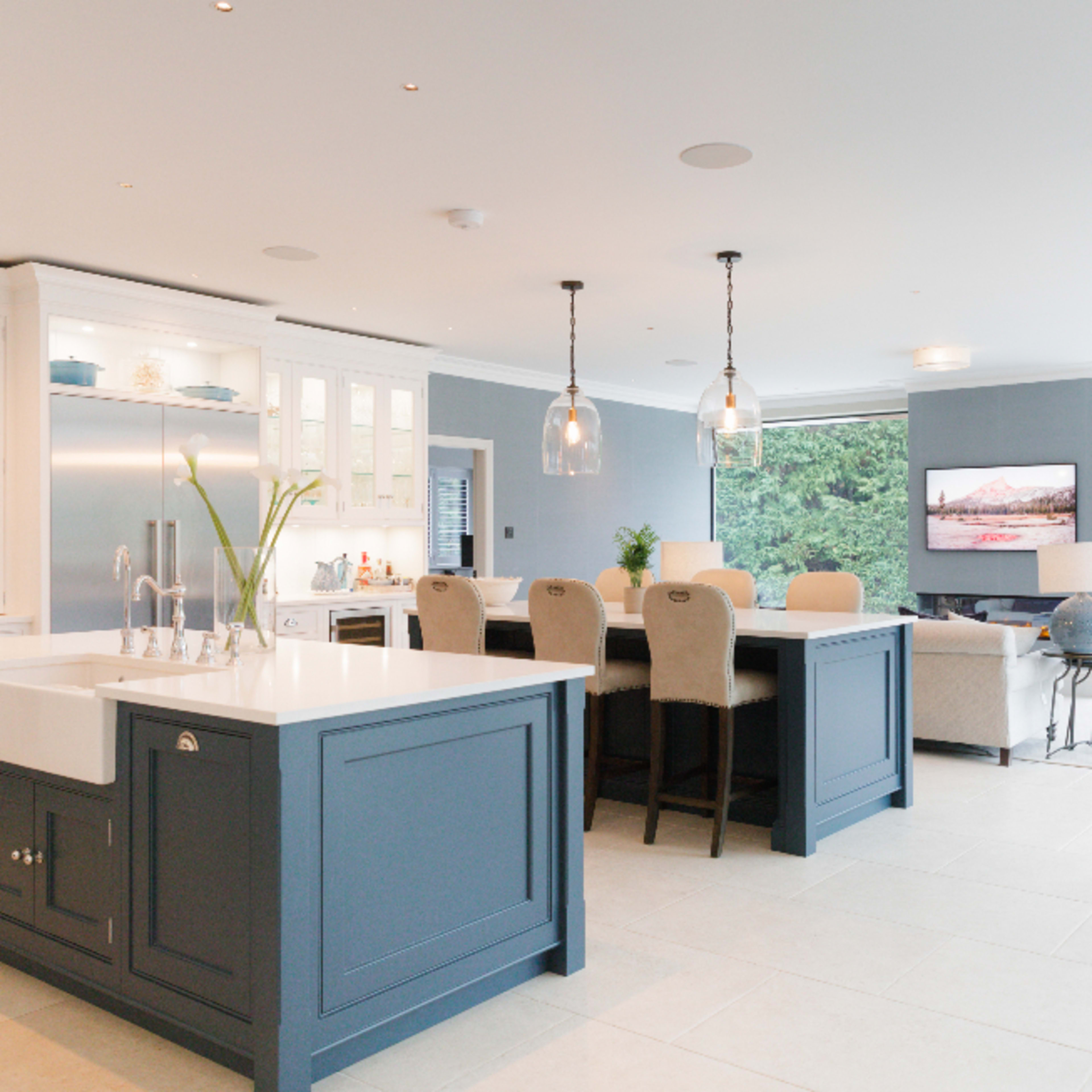 A modern kitchen features a central island with a sink, a dining area with beige chairs, and large windows overlooking a green outdoor area.
