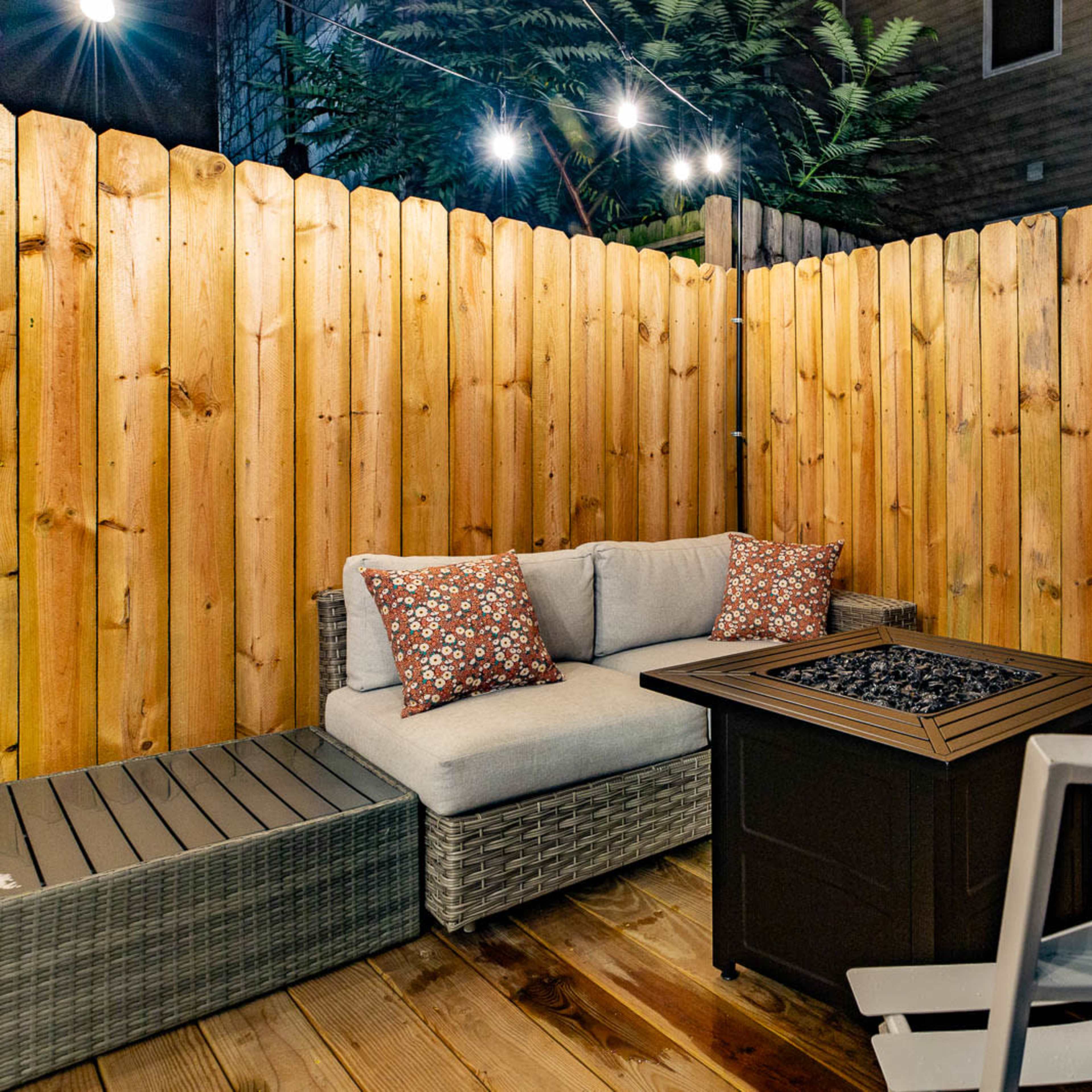 A cozy outdoor seating area features a fire pit surrounded by a sofa with decorative pillows and two white chairs, enclosed by a wooden fence.