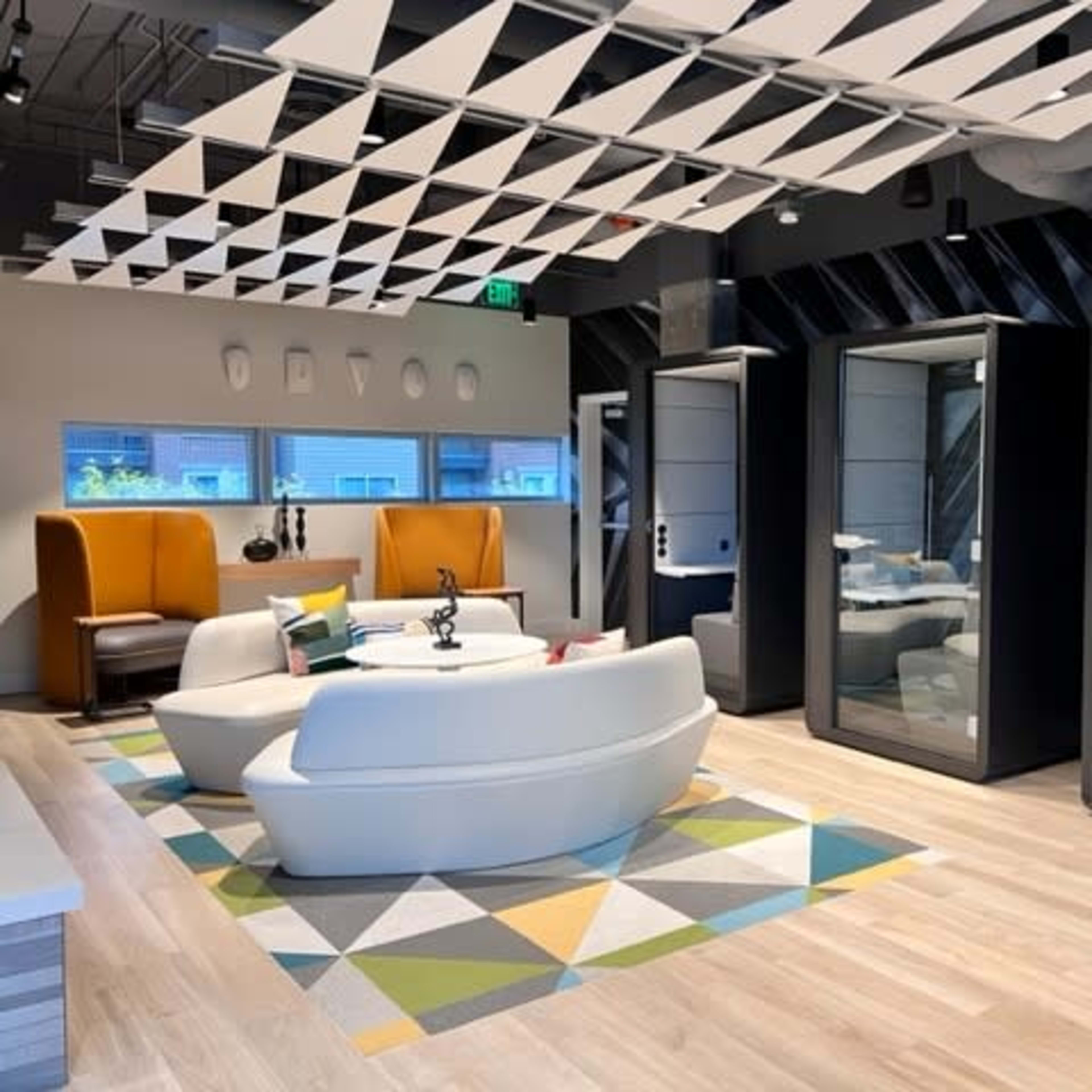 A modern office space featuring a seating area with a white sofa and colorful rug, along with glass-enclosed meeting pods on one side.