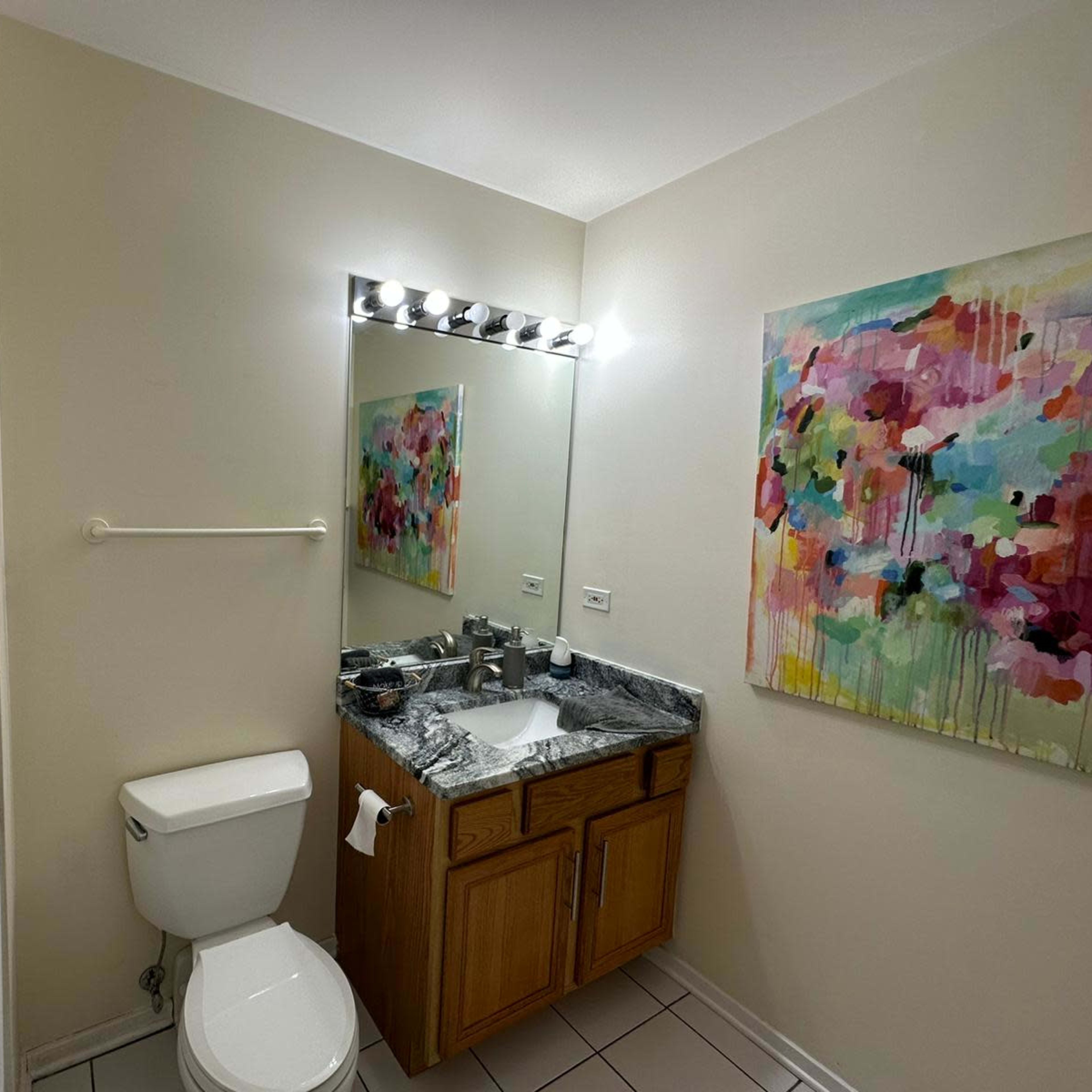 A small bathroom featuring a toilet, a sink with a granite countertop, a mirror with lighting, and a colorful abstract painting on the wall.