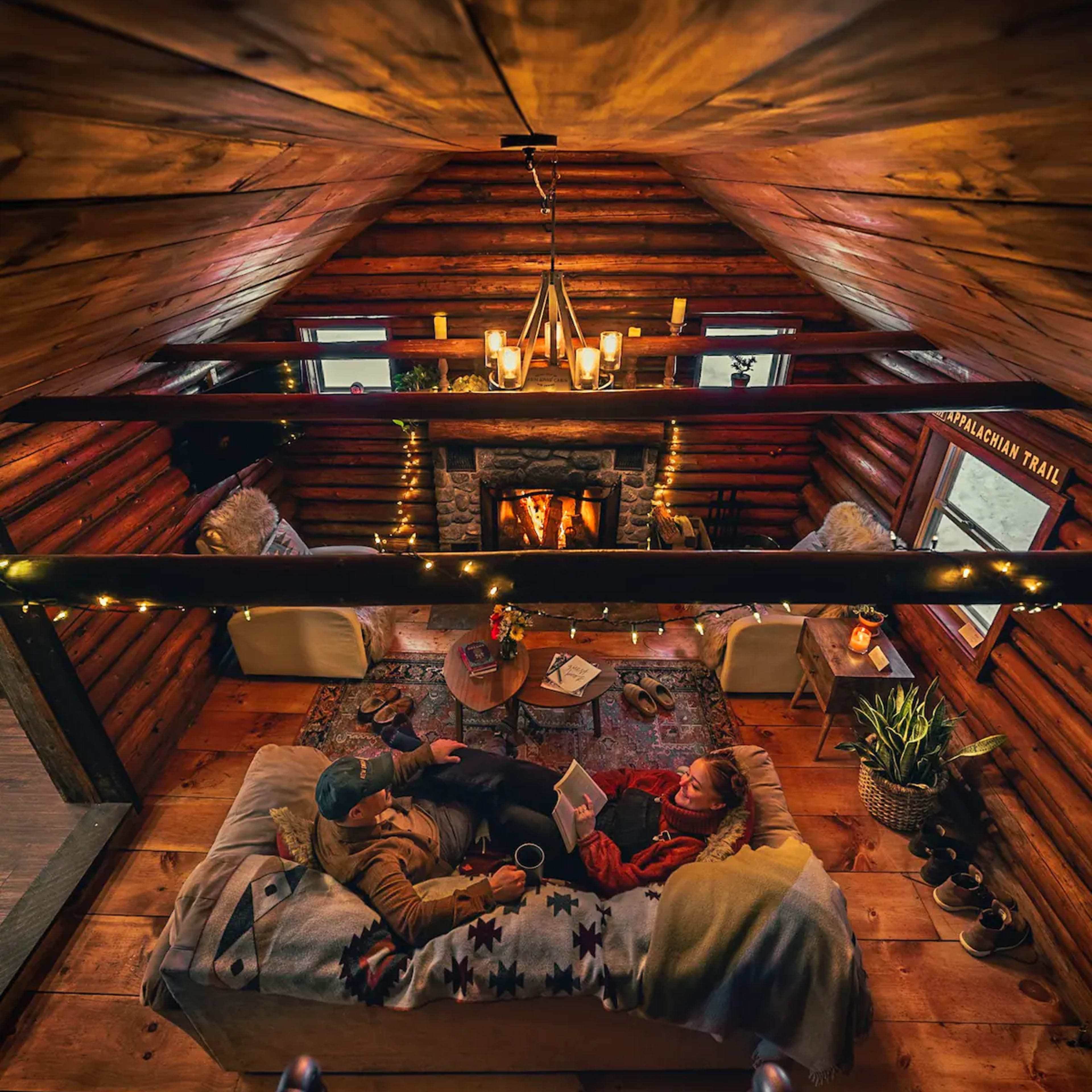 A couple relaxes on a cozy couch in a cabin, surrounded by wood-paneled walls and a glowing fireplace, with warm lighting and plants nearby.