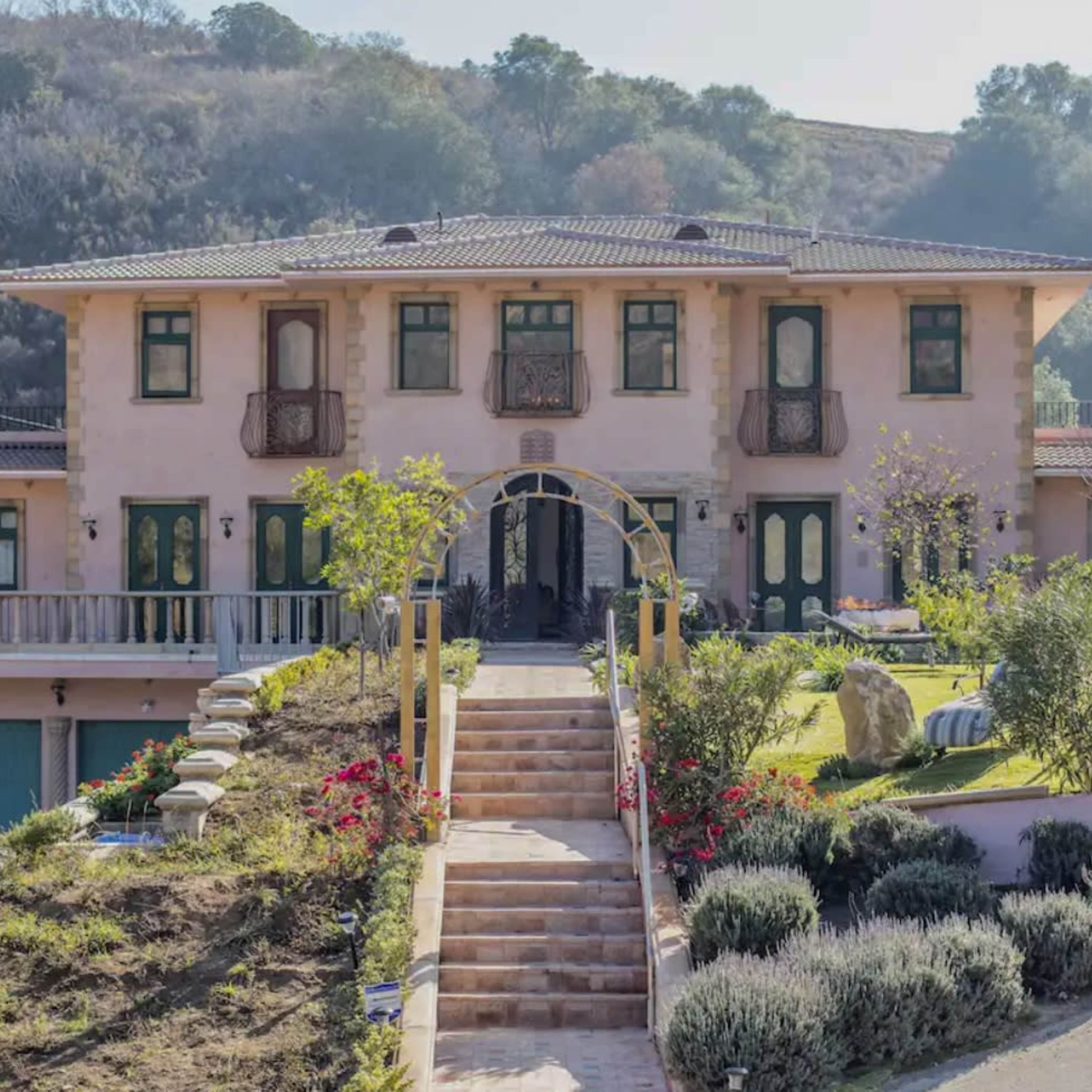 A large, two-story house with a landscaped front yard features a prominent staircase leading to the entrance.