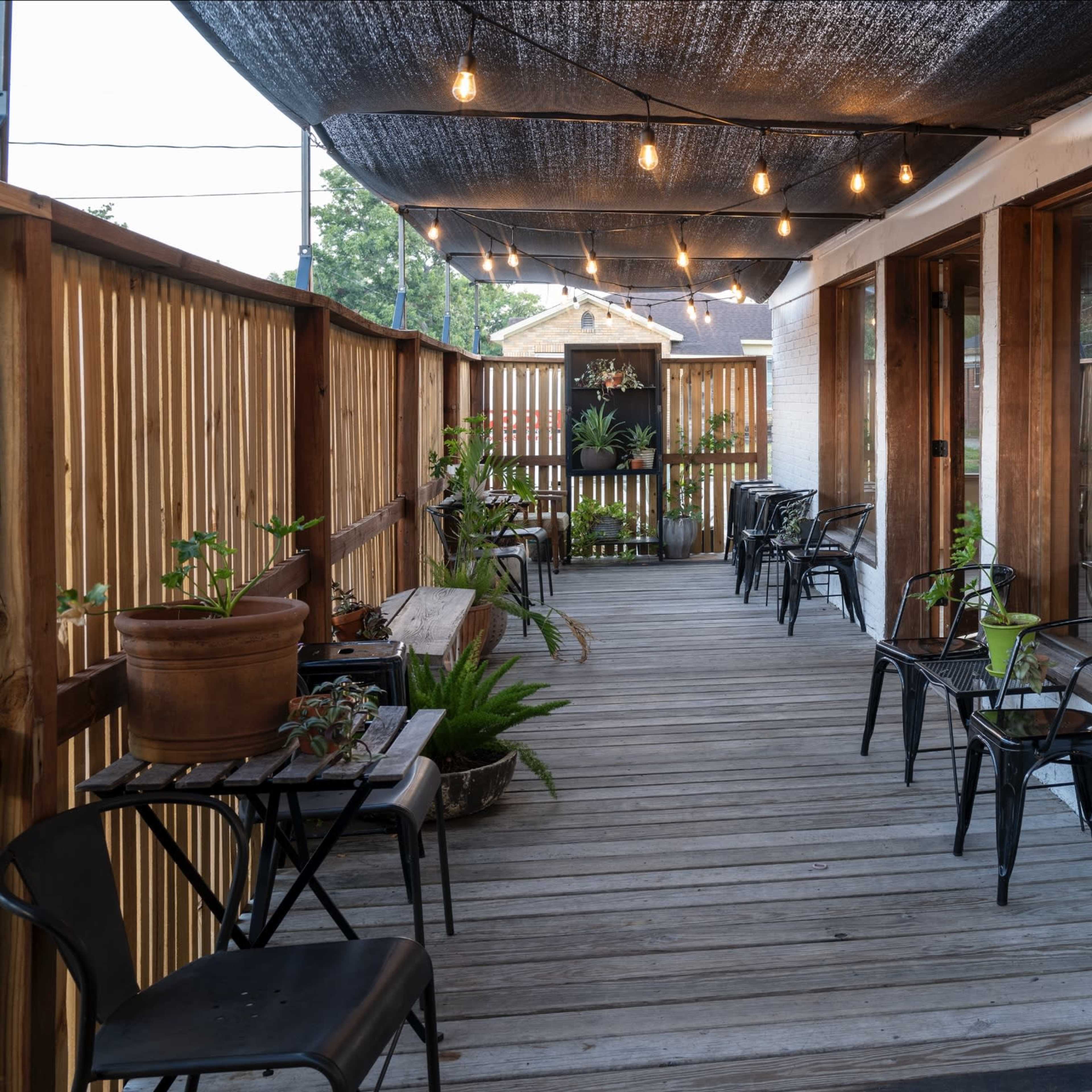 A wooden deck with potted plants and empty chairs under decorative string lights and a canopy.