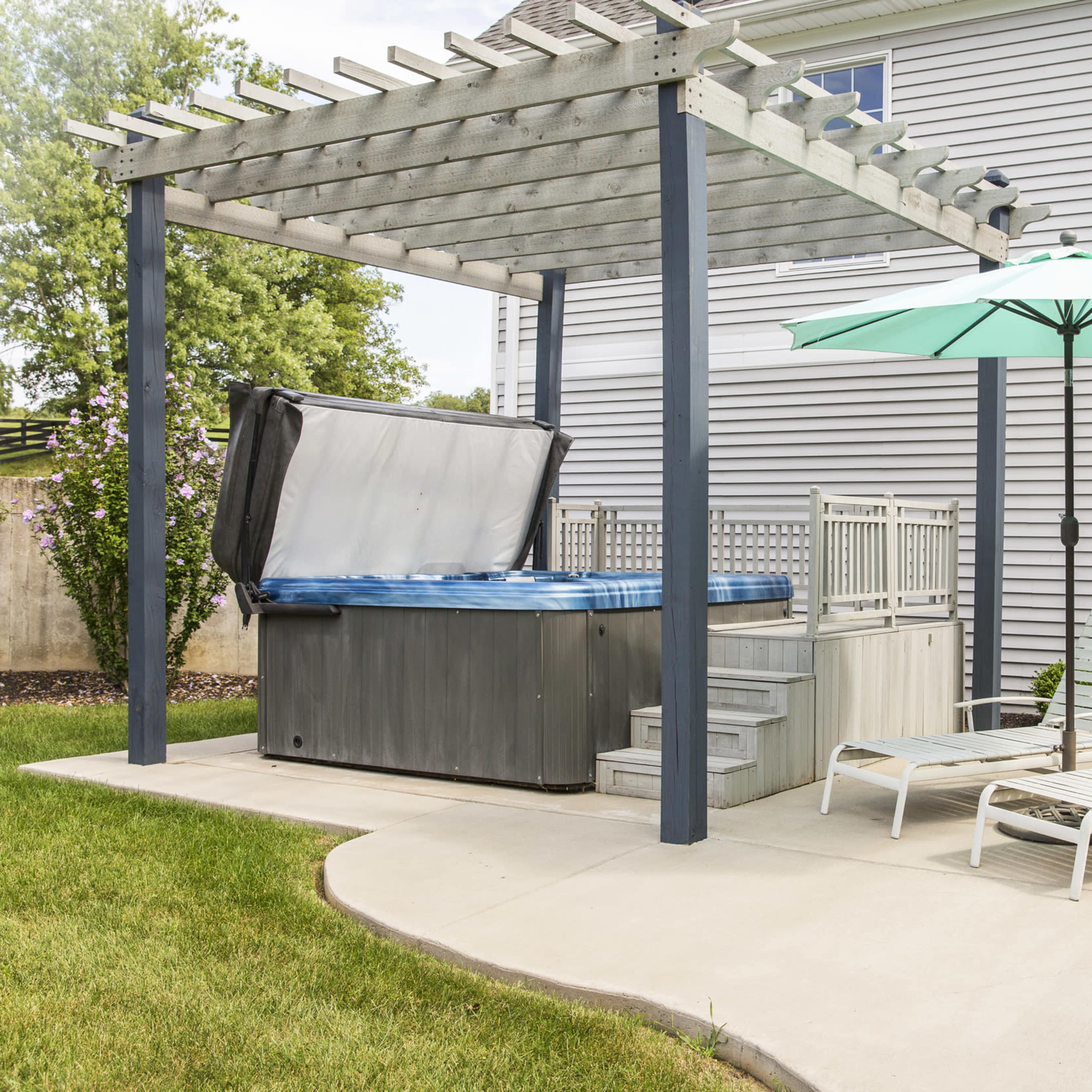 A hot tub is placed under a wooden pergola beside a patio featuring a green umbrella and lounge chairs.