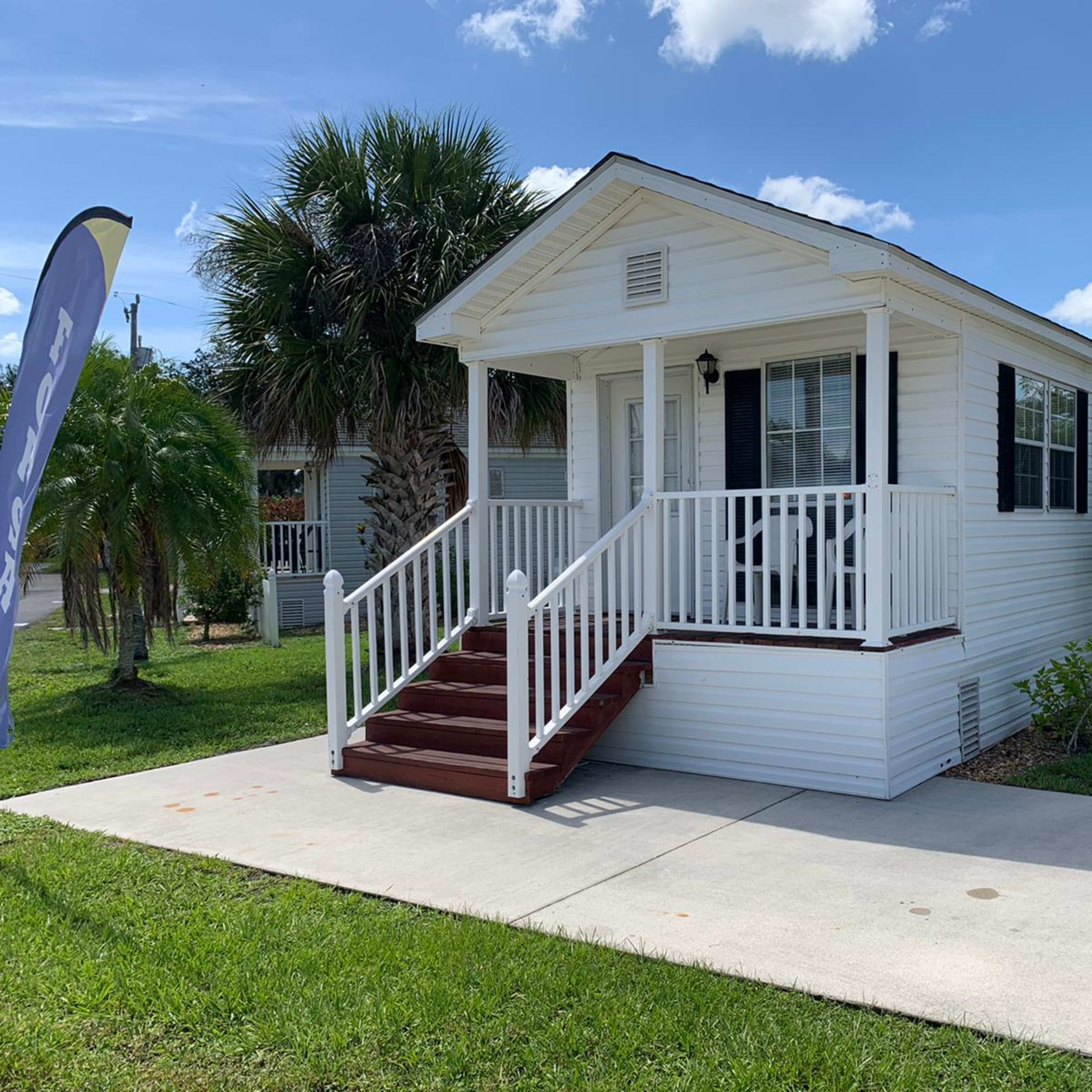 A white, single-story house with a porch and steps, situated on a grassy lawn with palm trees in the background.