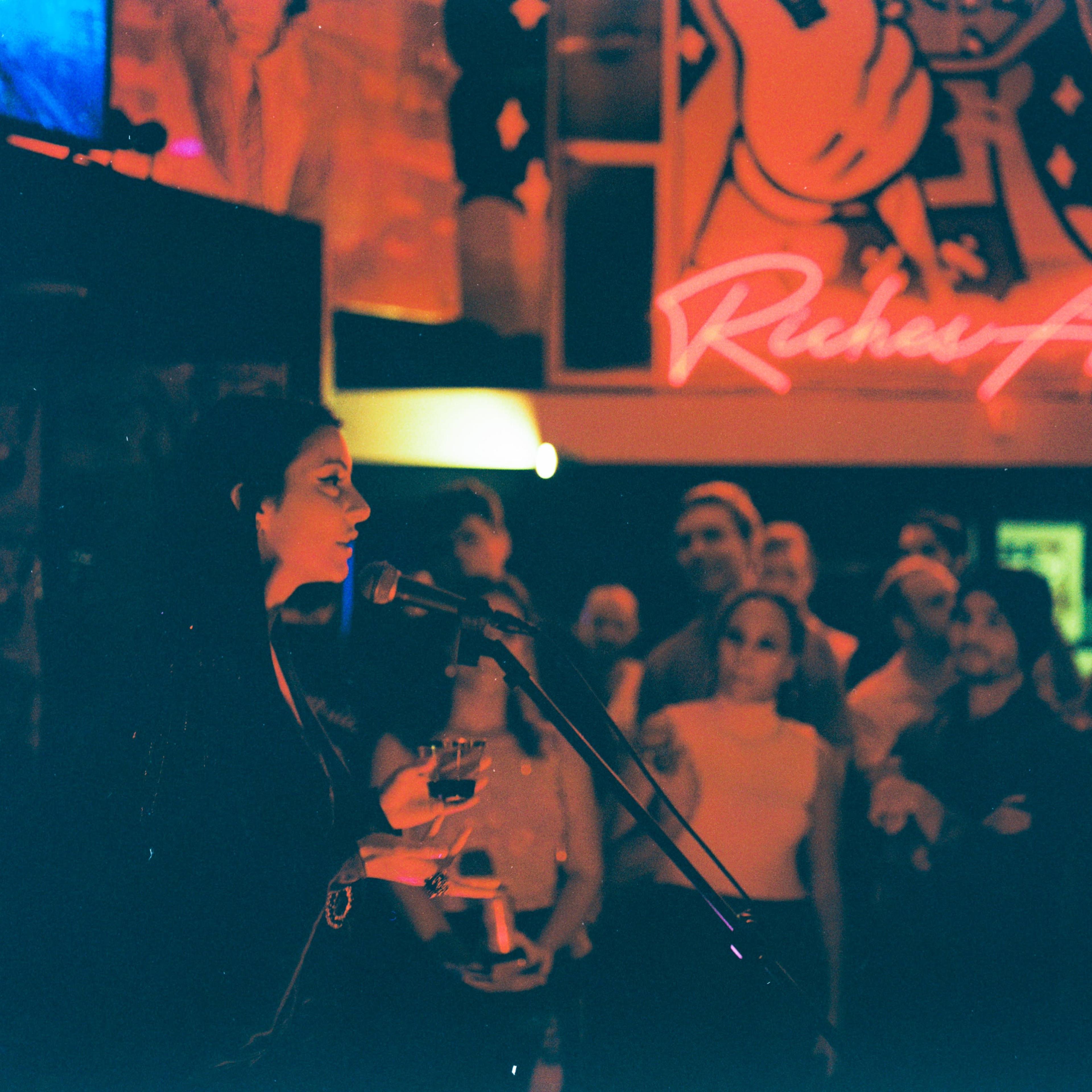 A woman speaking into a microphone holds a drink while a crowd listens in an art gallery featuring vibrant decorations.