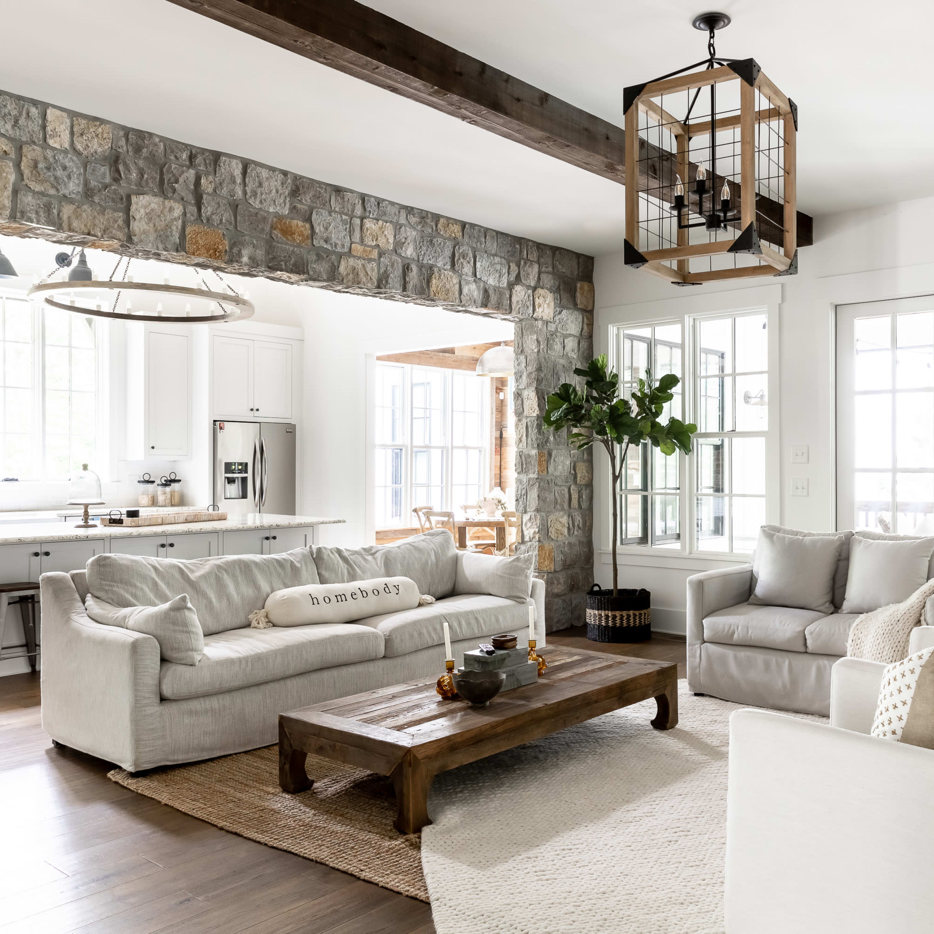 A modern living room with two sofas facing a wooden coffee table, set against a stone wall, leading into a kitchen area with white cabinetry and large windows.