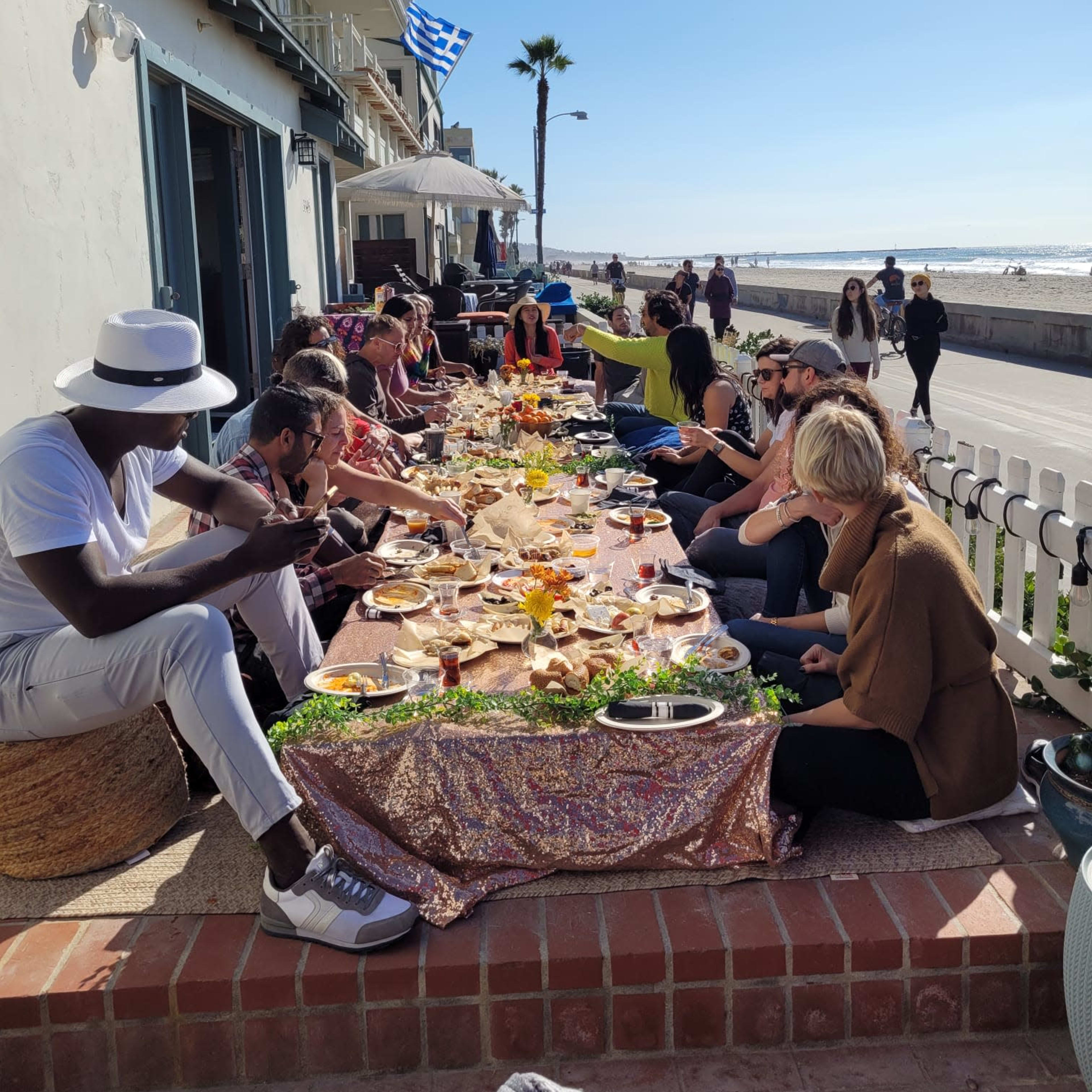 A long table is set with a variety of food and drinks along a beachfront patio where people are seated and enjoying their meal under a clear blue sky.