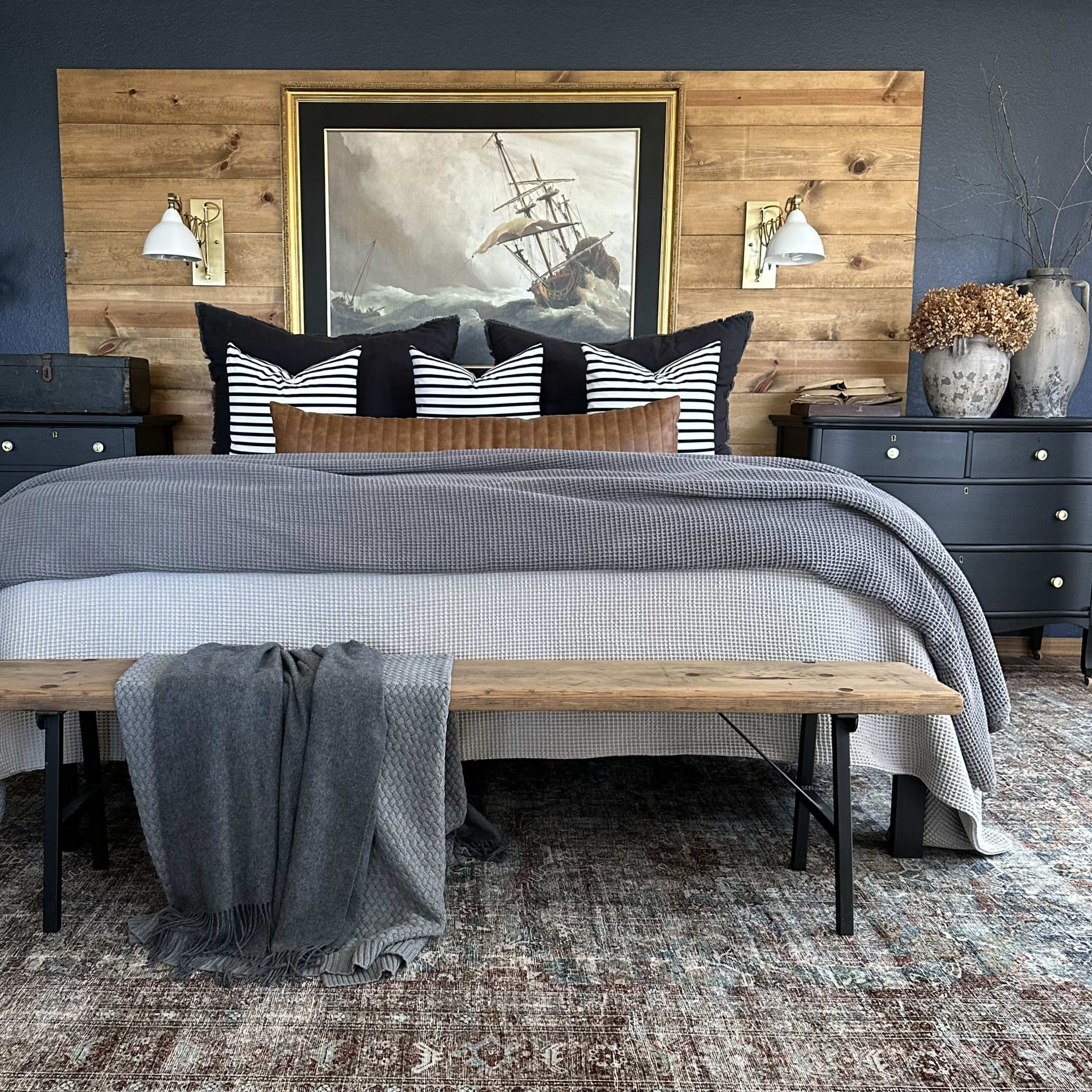 A neatly arranged bedroom featuring a large bed with a gray blanket, striped pillows, a wooden bench at the foot, and decorative elements against a wooden accent wall.
