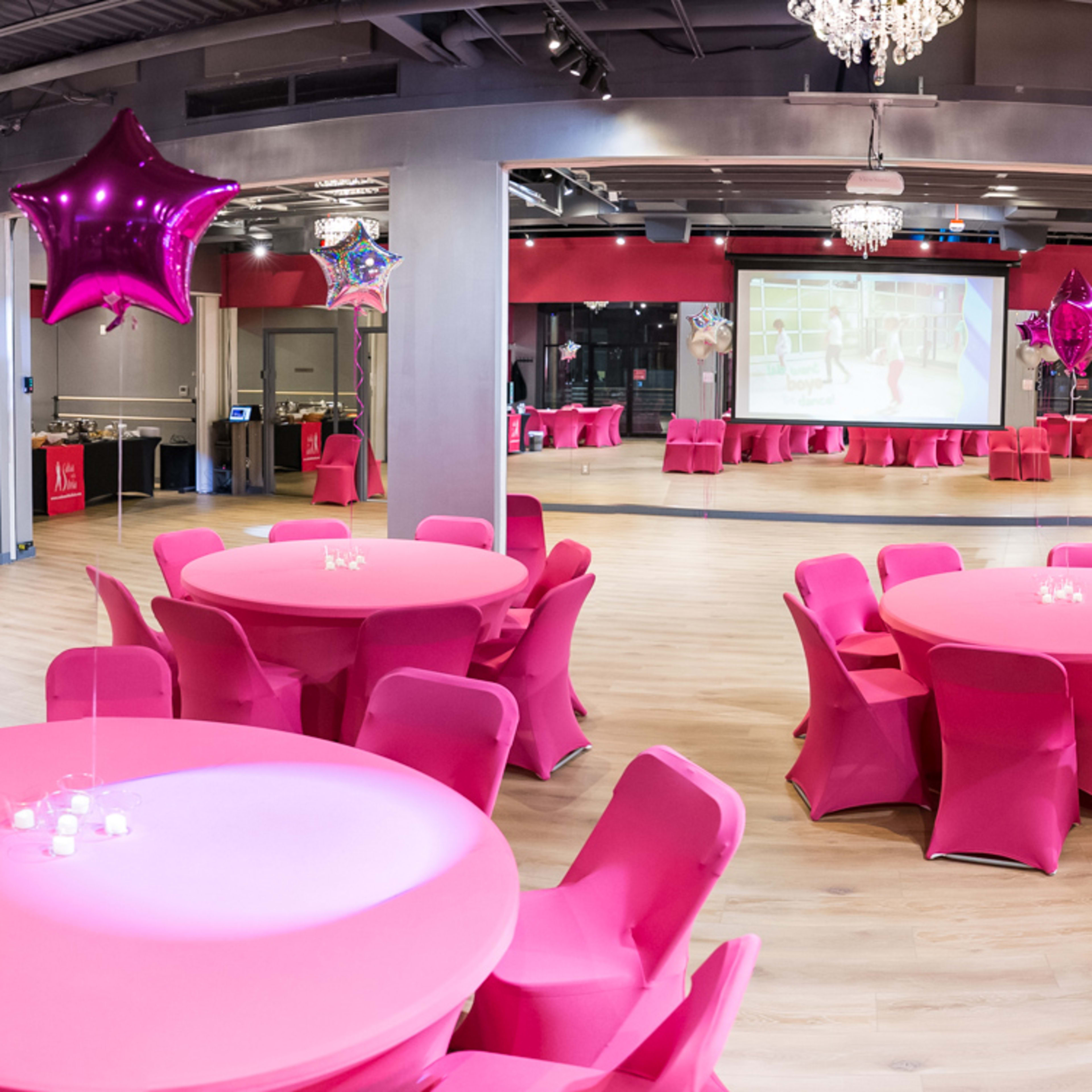 A spacious event hall features round pink tables covered with pink tablecloths and adorned with small candles, surrounded by pink walls and decorative star balloons.