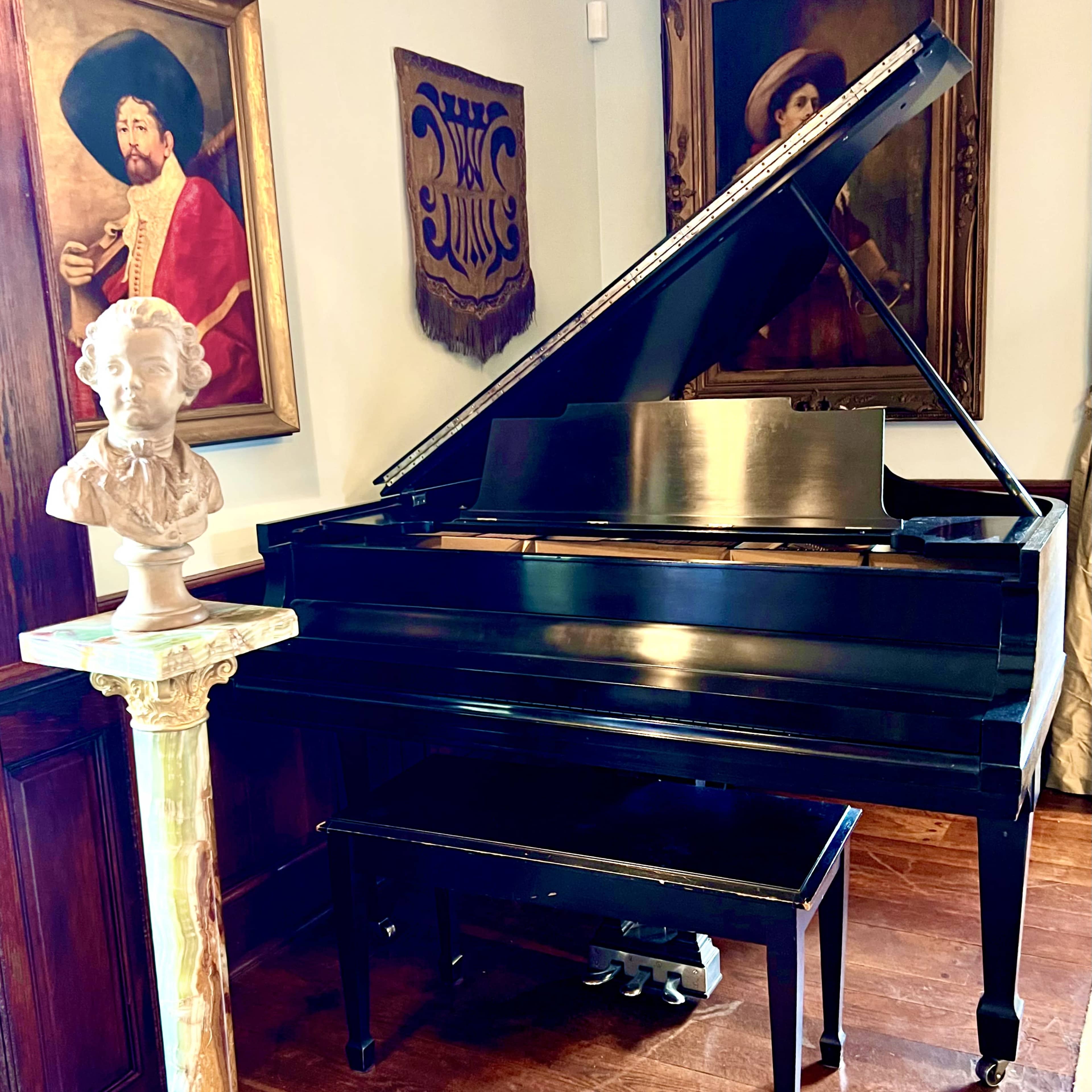 A black grand piano is positioned beside a white marble bust on a gold-painted pedestal, with framed portraits hanging on the walls.