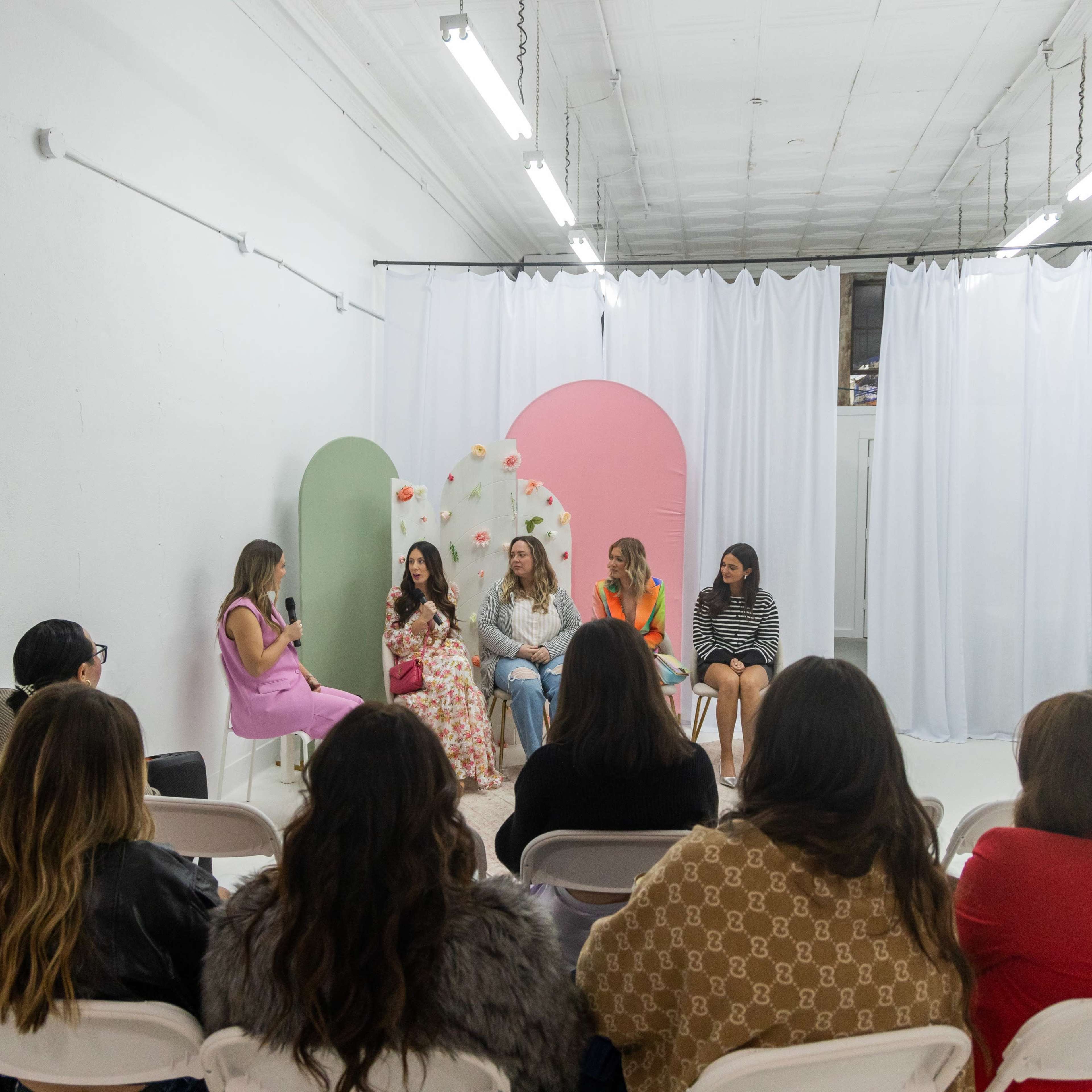 A group of women sits on chairs in a minimalist space, participating in a panel discussion while an audience faces them.