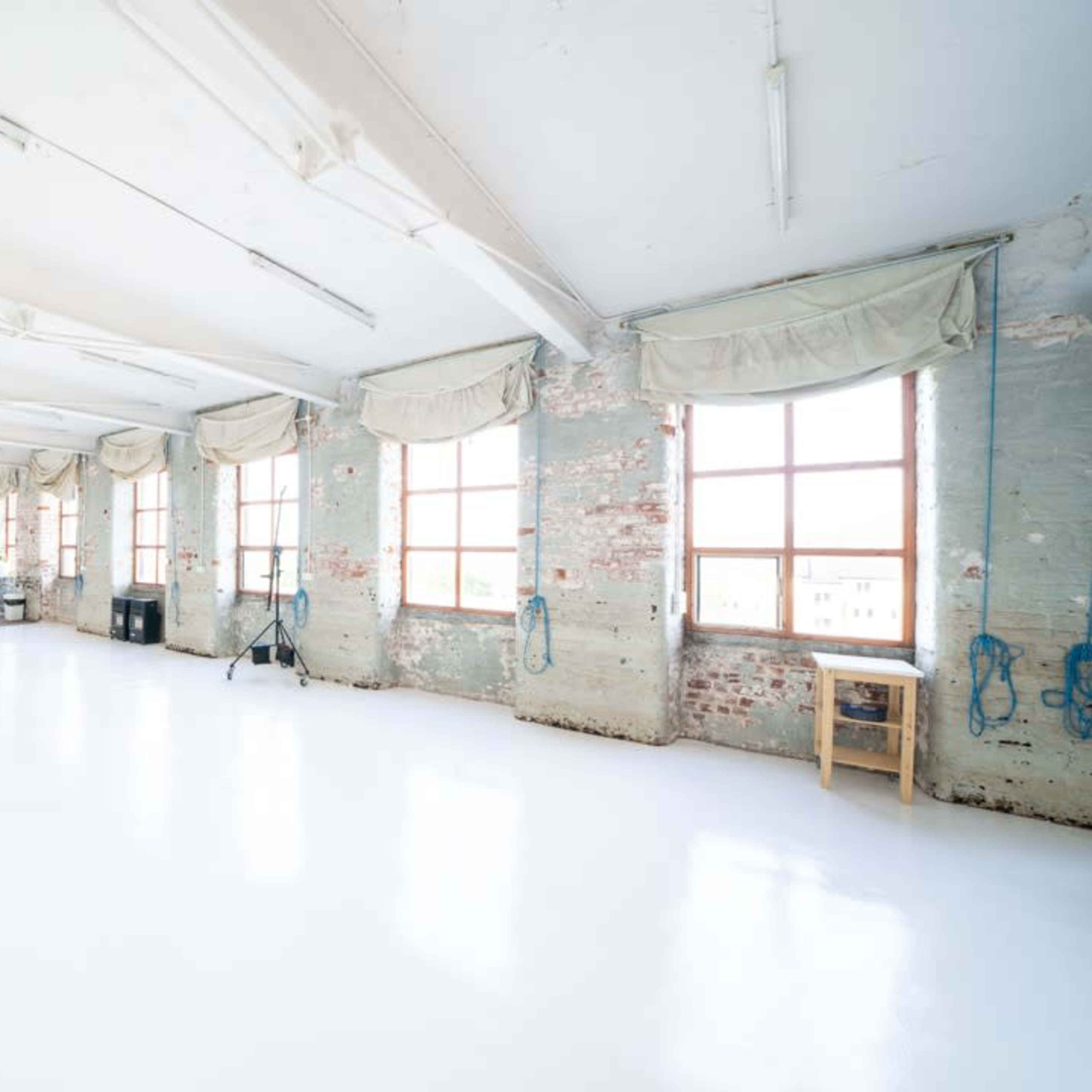 A spacious, well-lit studio with large windows and a polished white floor, featuring exposed brick walls and minimal furnishings.