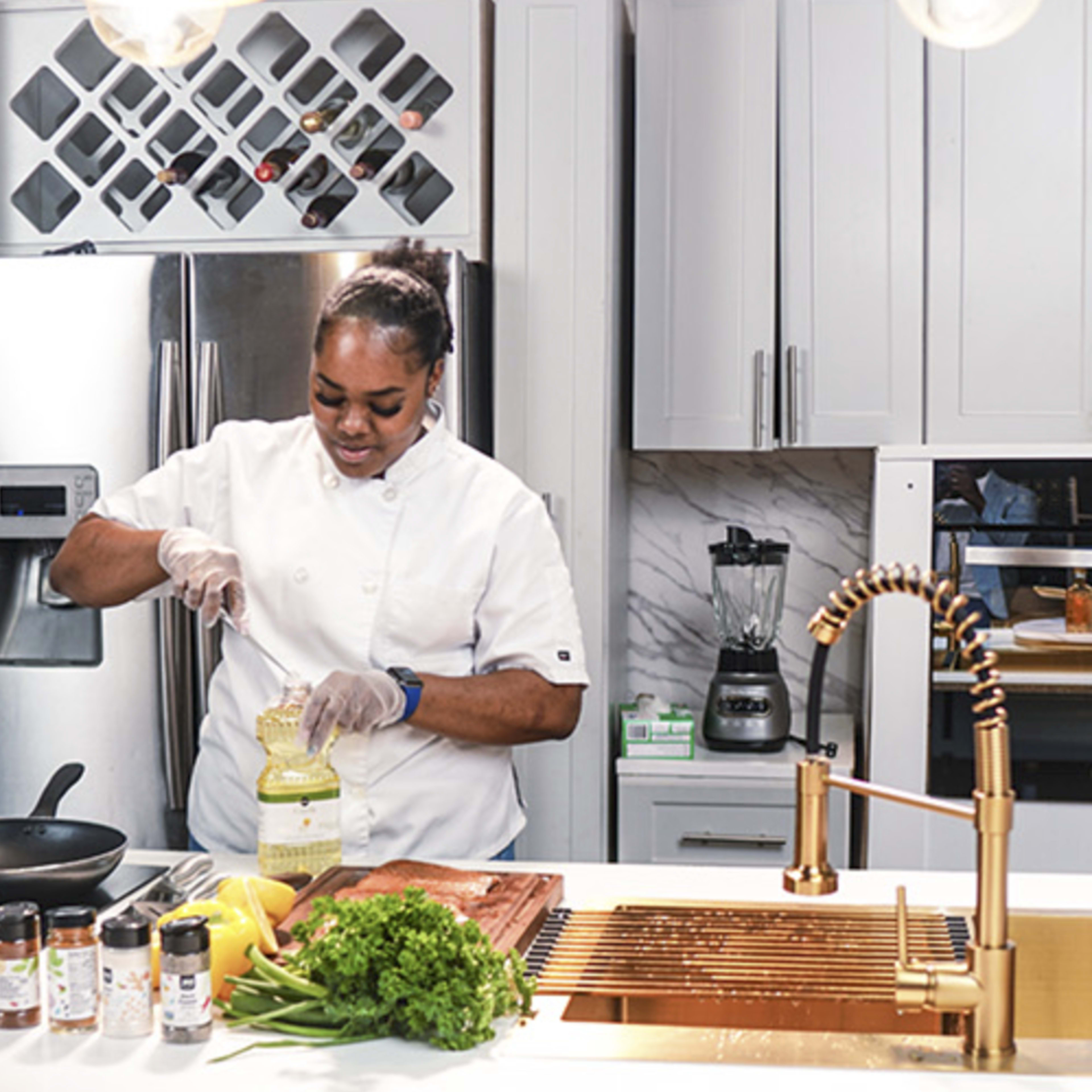 A chef wearing a white uniform is pouring oil into a frying pan in a modern kitchen equipped with stainless steel appliances.