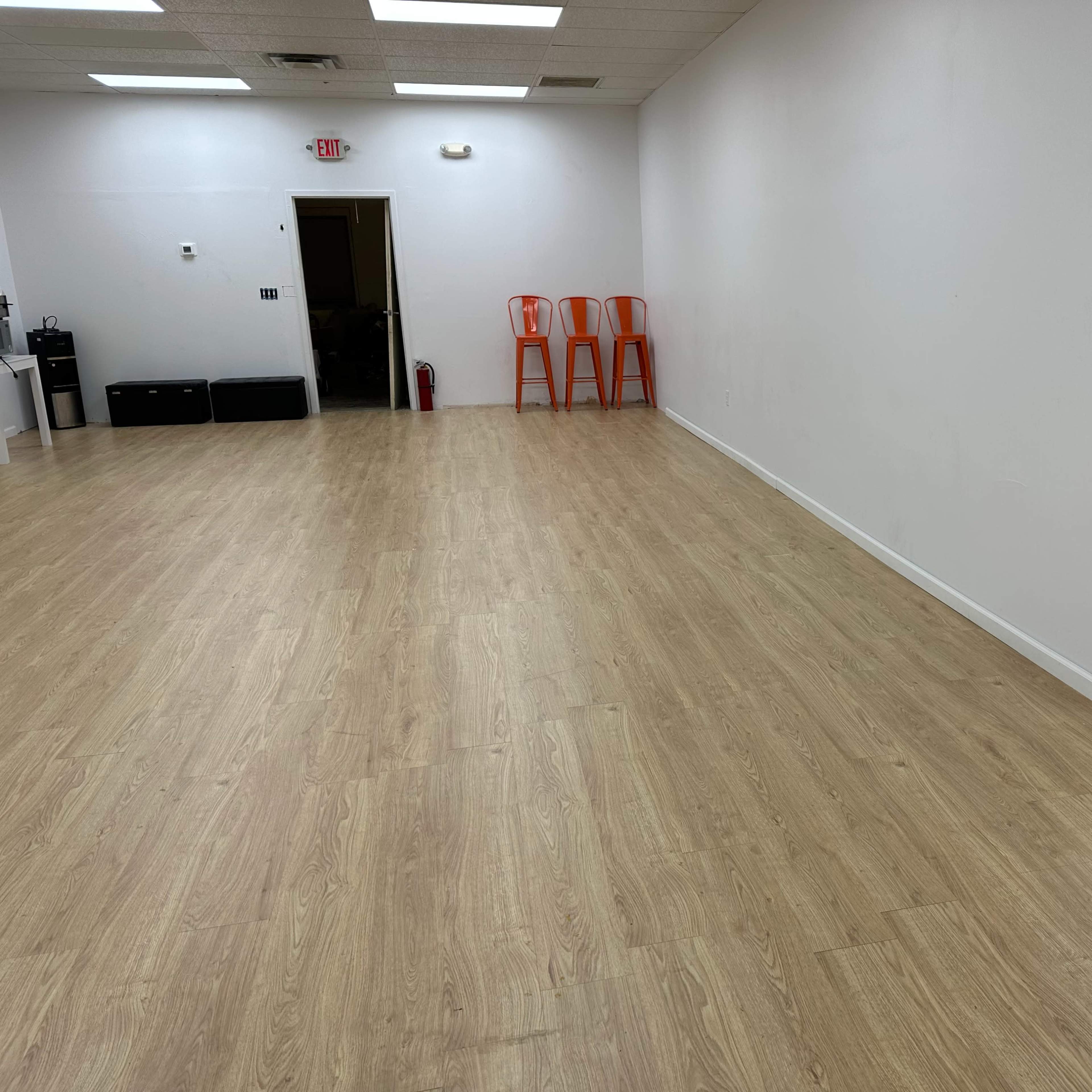 An empty room with light wooden flooring, white walls, three orange chairs stacked in the corner, and a mirror on one wall.