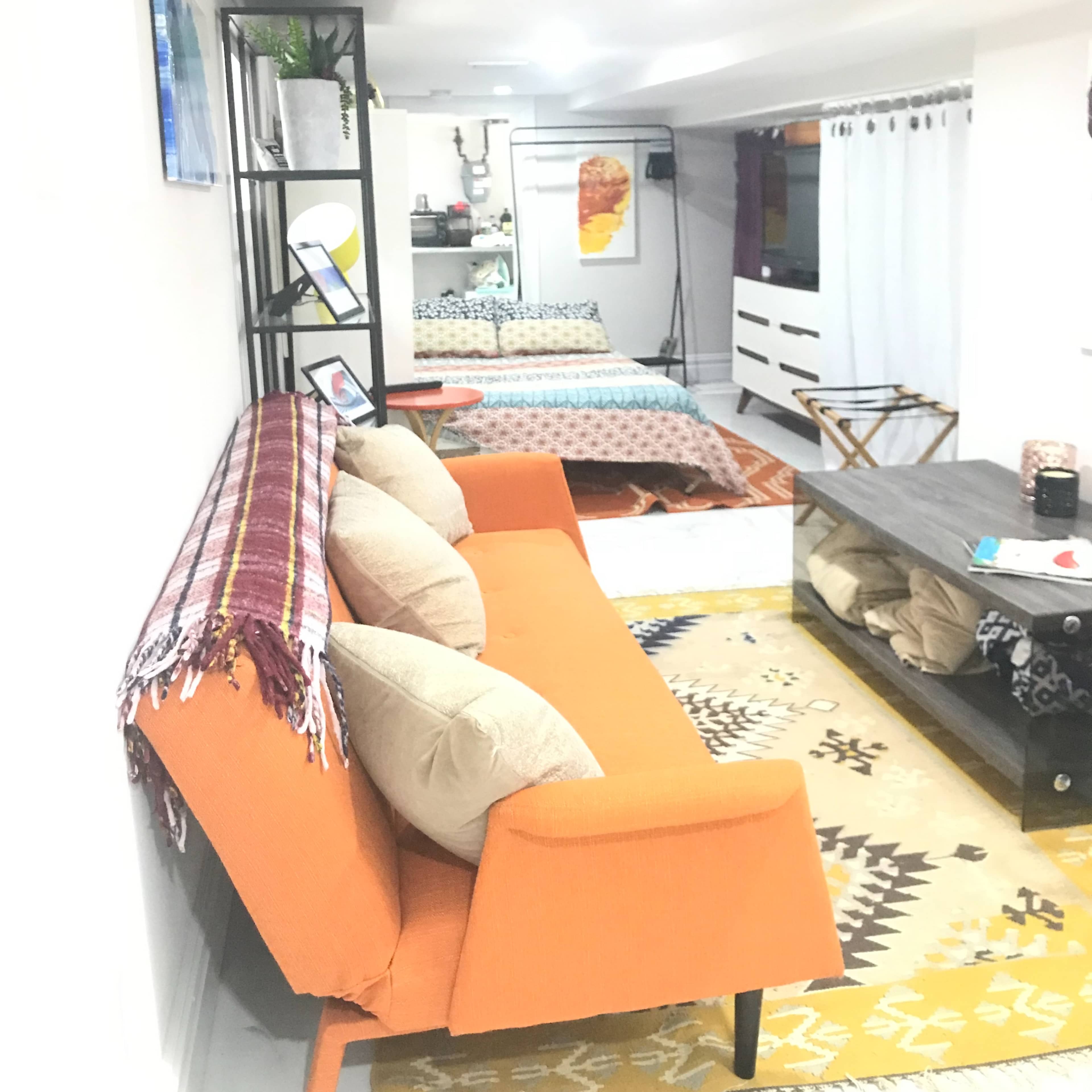A cozy interior space featuring an orange sofa, decorative pillows, a coffee table, and a bedroom area with a patterned bedspread visible in the background.
