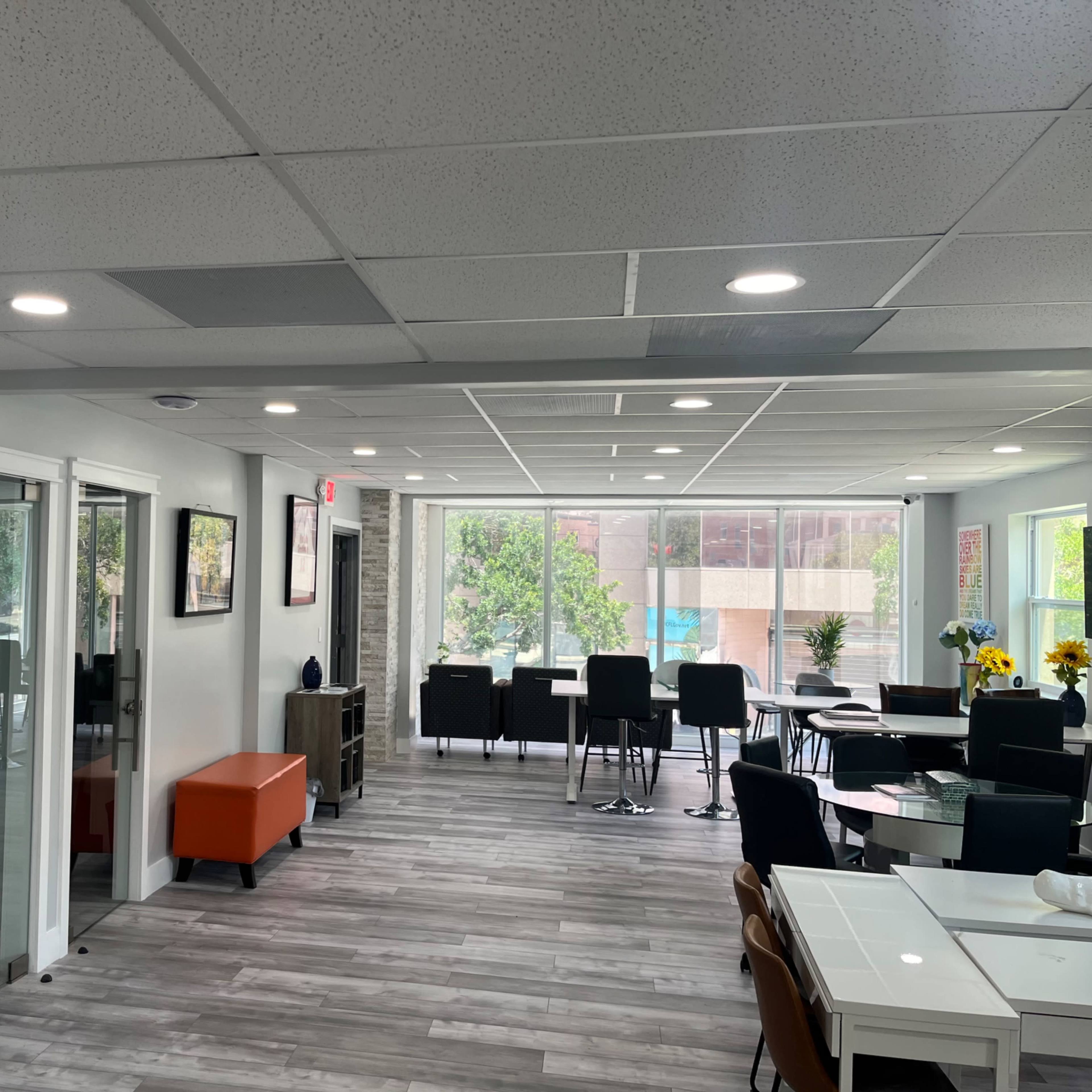 A modern office space with a reception area, tables, chairs, and large windows allowing natural light to enter.