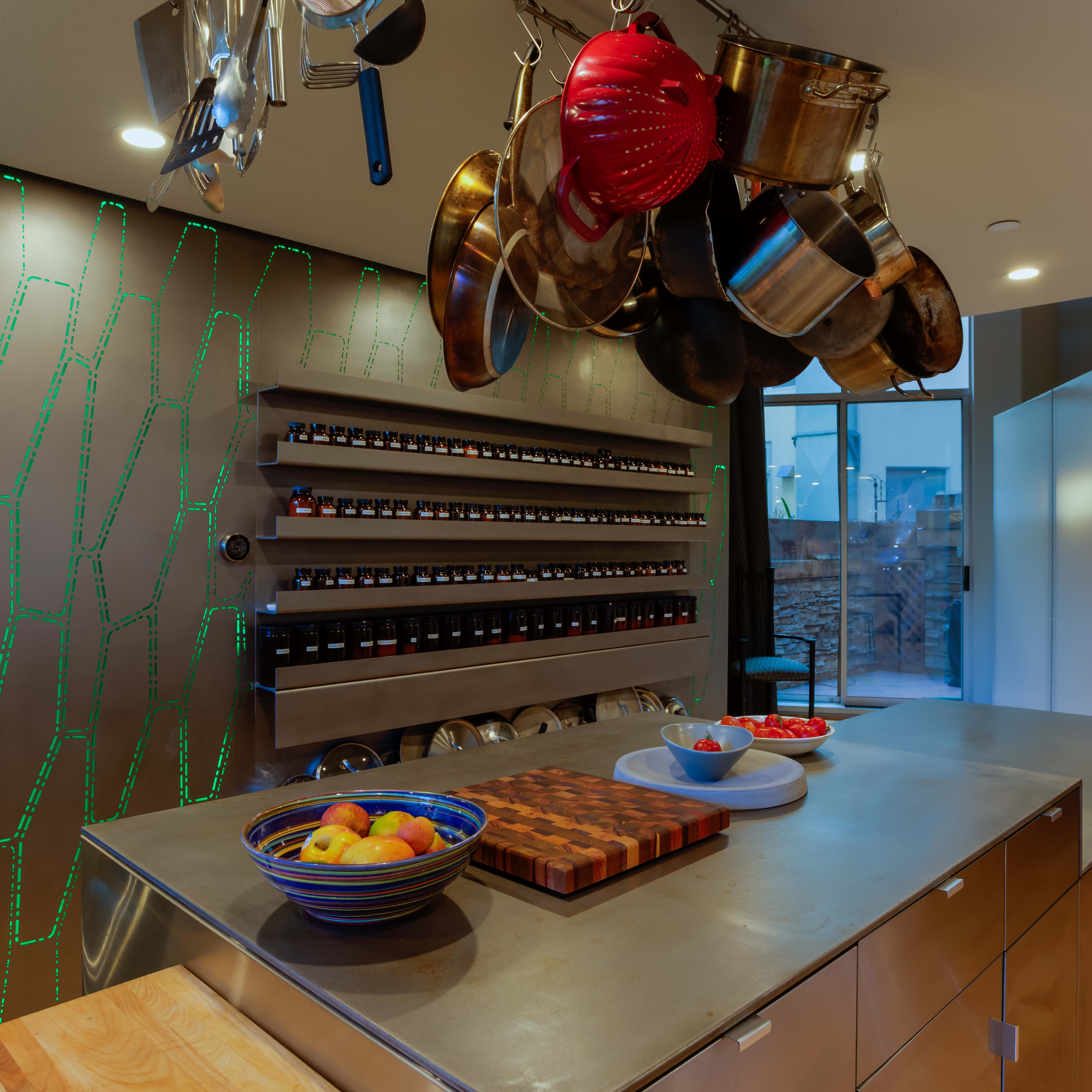 A modern kitchen features a central island with a wooden cutting board, bowls of fruit, and a rack of spices mounted on the wall, with hanging pots above.