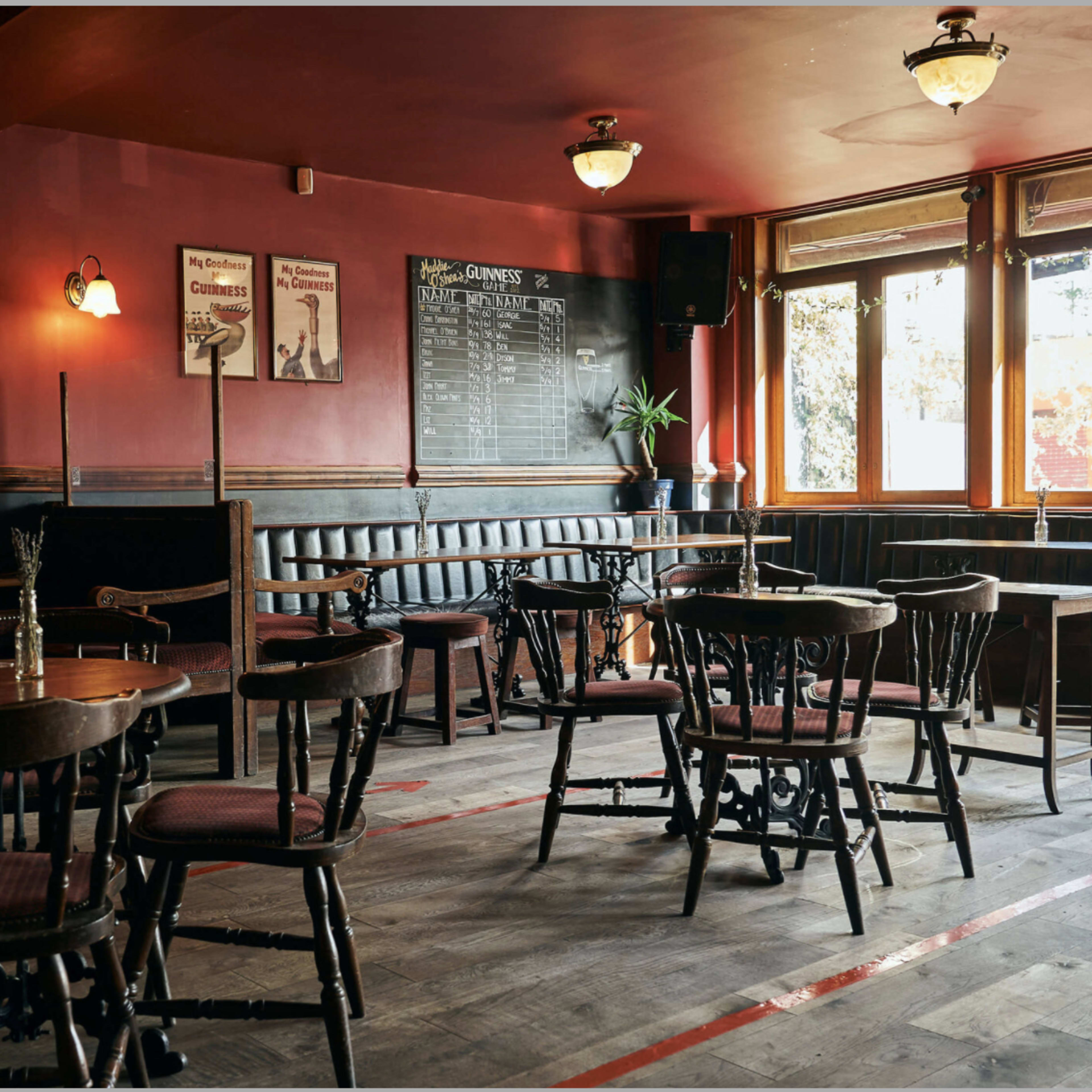 A vintage-style pub interior with wooden tables and chairs arranged around the space, decorative mirrors on the walls, and a chalkboard menu near the large windows.