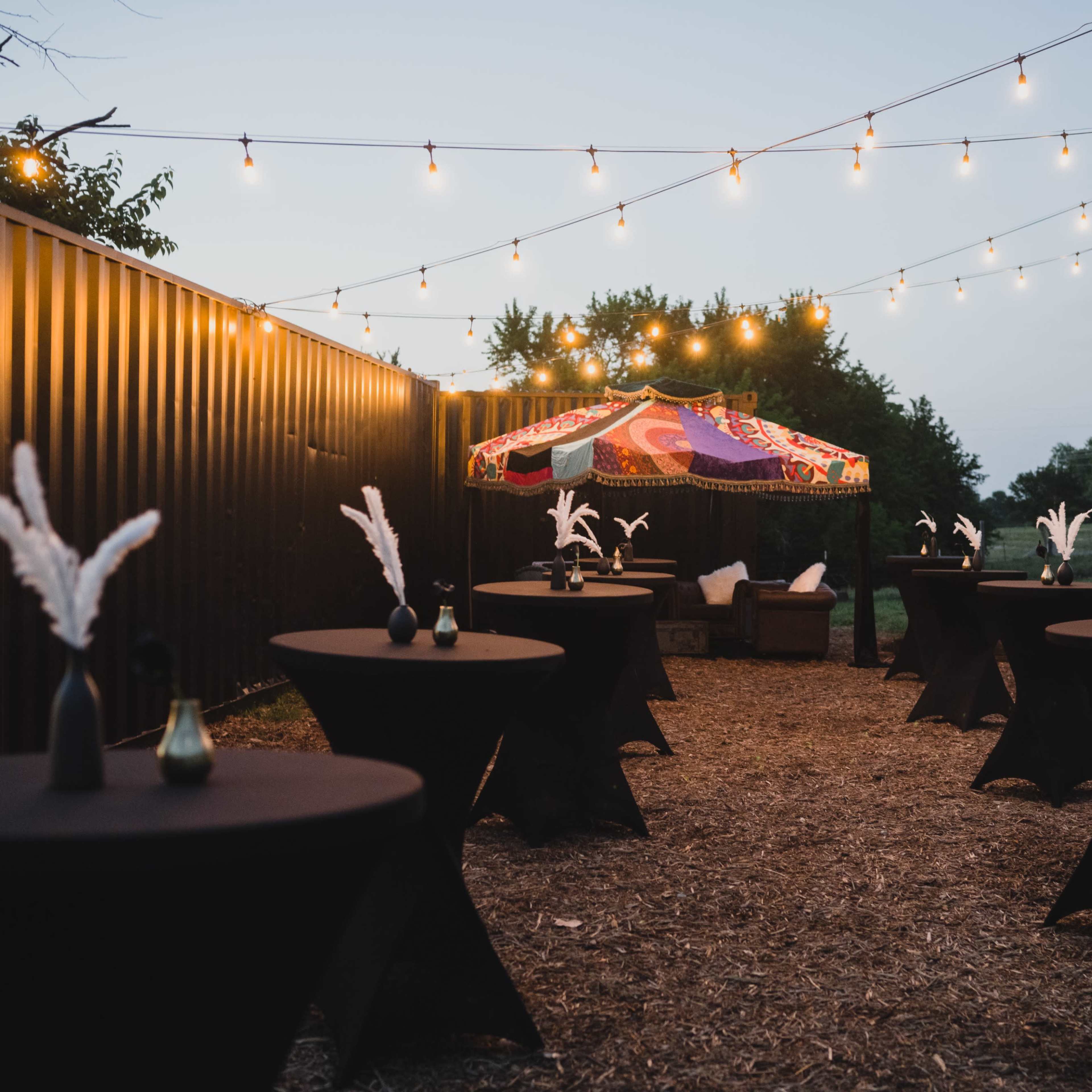 The scene features an outdoor event space with string lights overhead, black cocktail tables adorned with decorative feathers, and a colorful tent in the background.