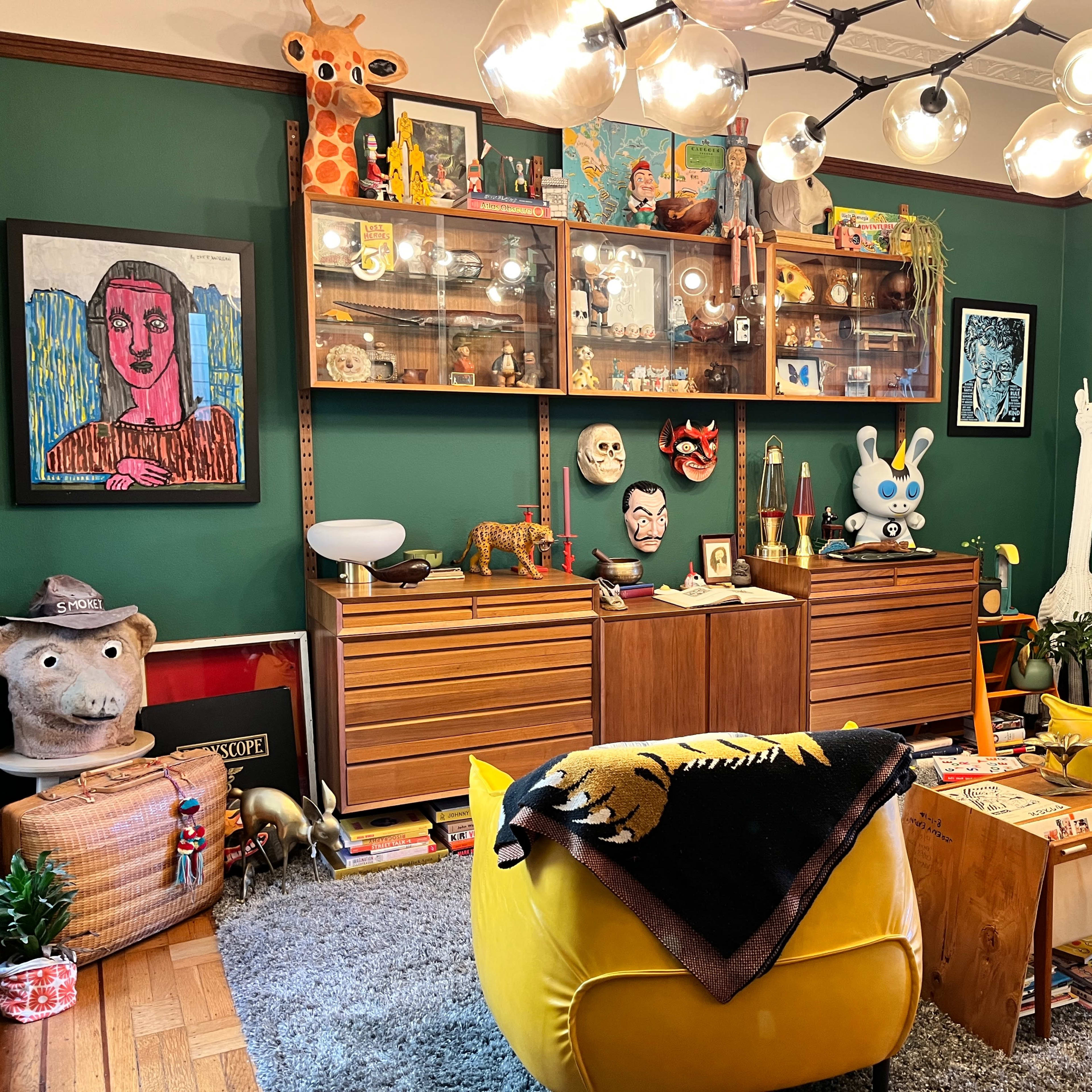 A colorful living room with yellow armchairs, a green wall, and a wooden cabinet displaying various art pieces and collectibles.