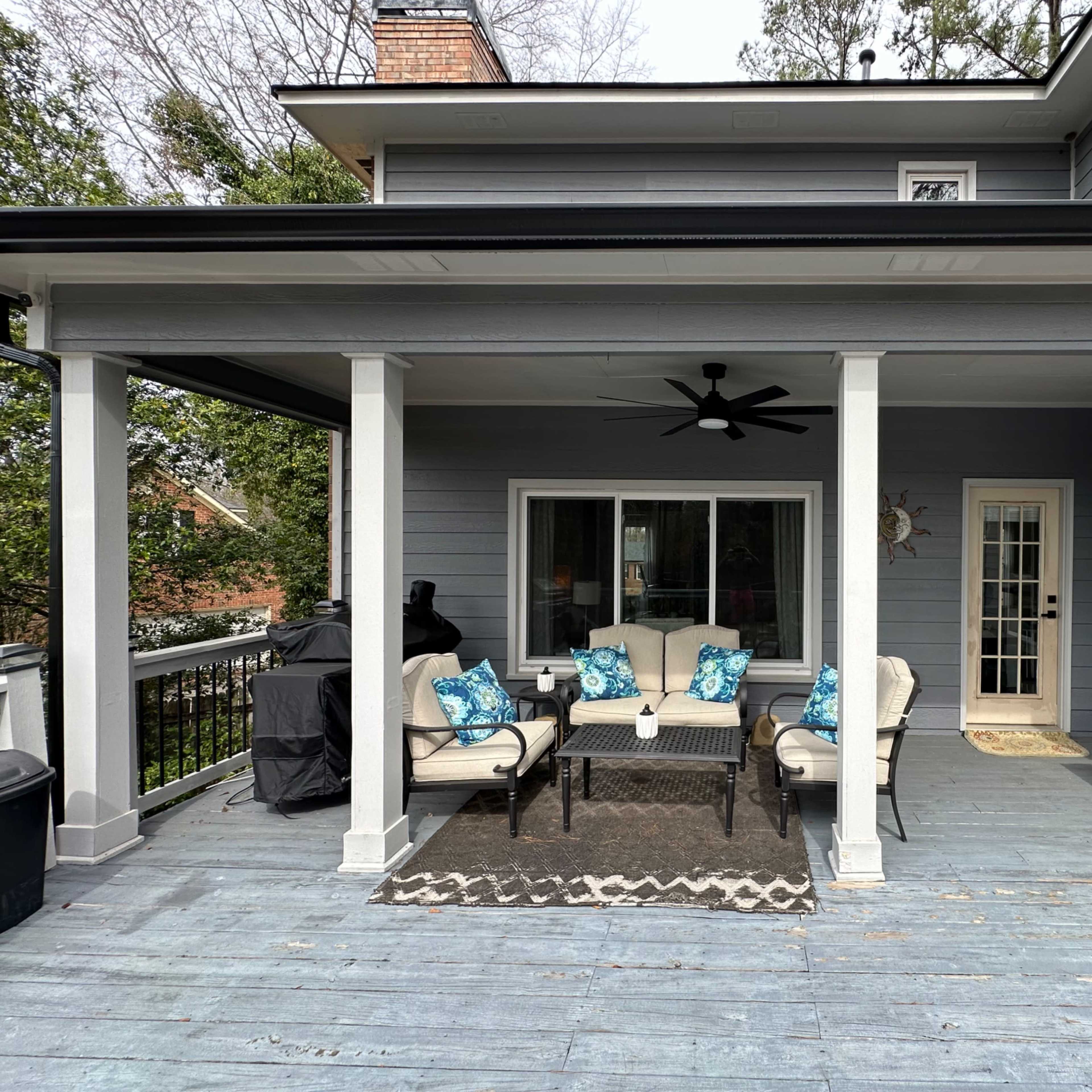 A covered porch with a seating area consisting of beige chairs with blue decorative pillows, a small table, and a door leading into the house.