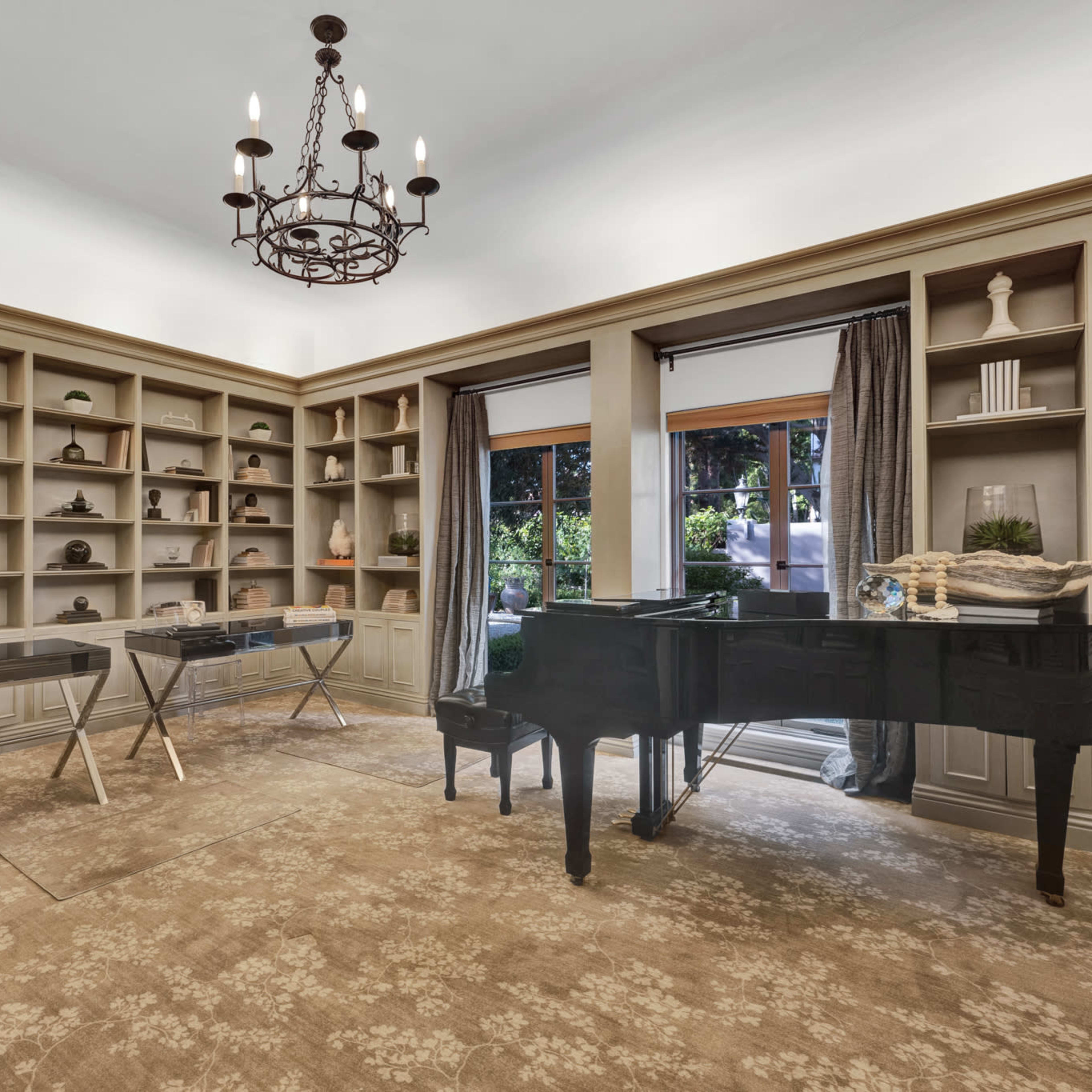 A well-furnished study with a grand piano, two glass desks, and a large bookcase filled with decor items, complemented by windows that provide natural light.