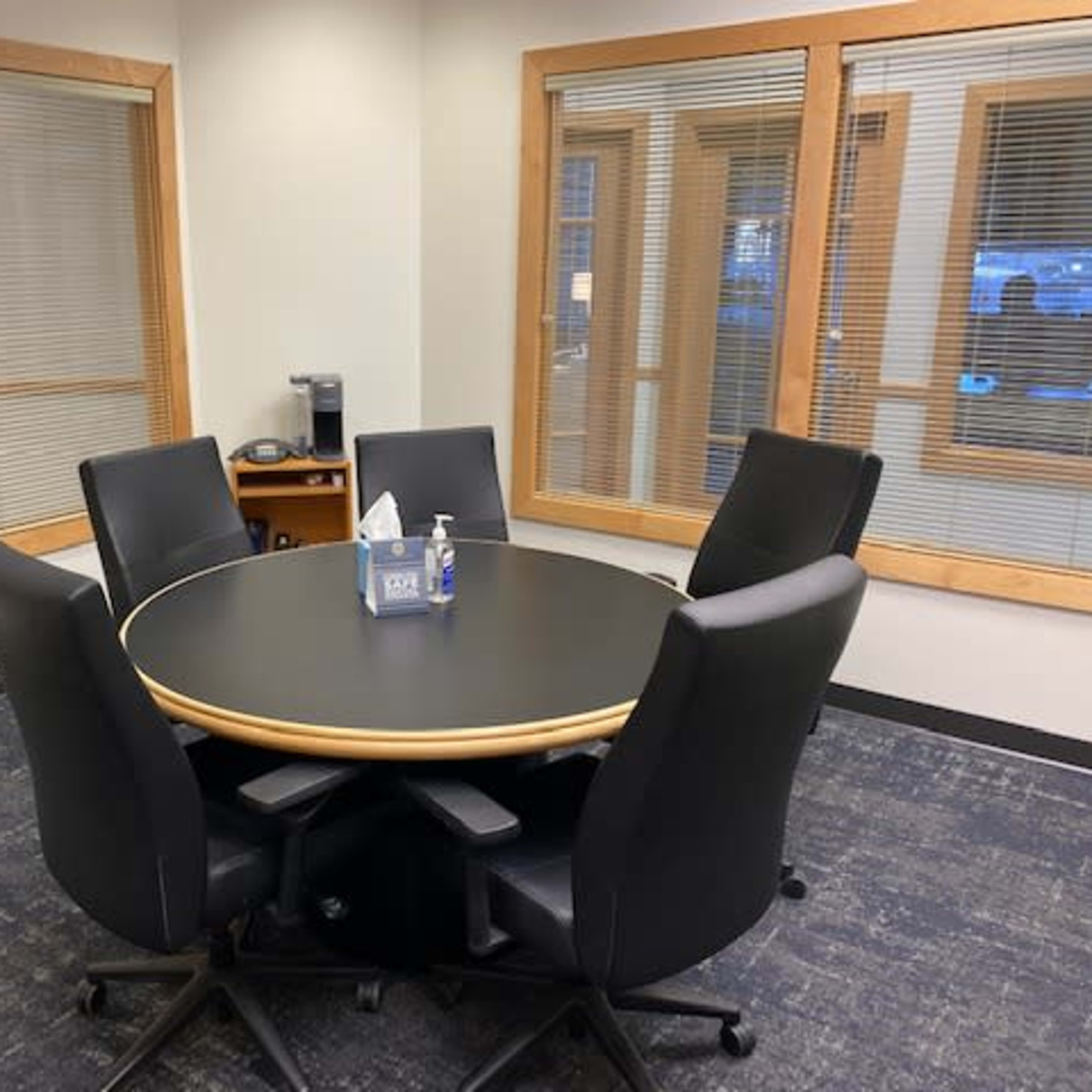 A round black conference table surrounded by five black office chairs in a well-lit meeting room with large windows.