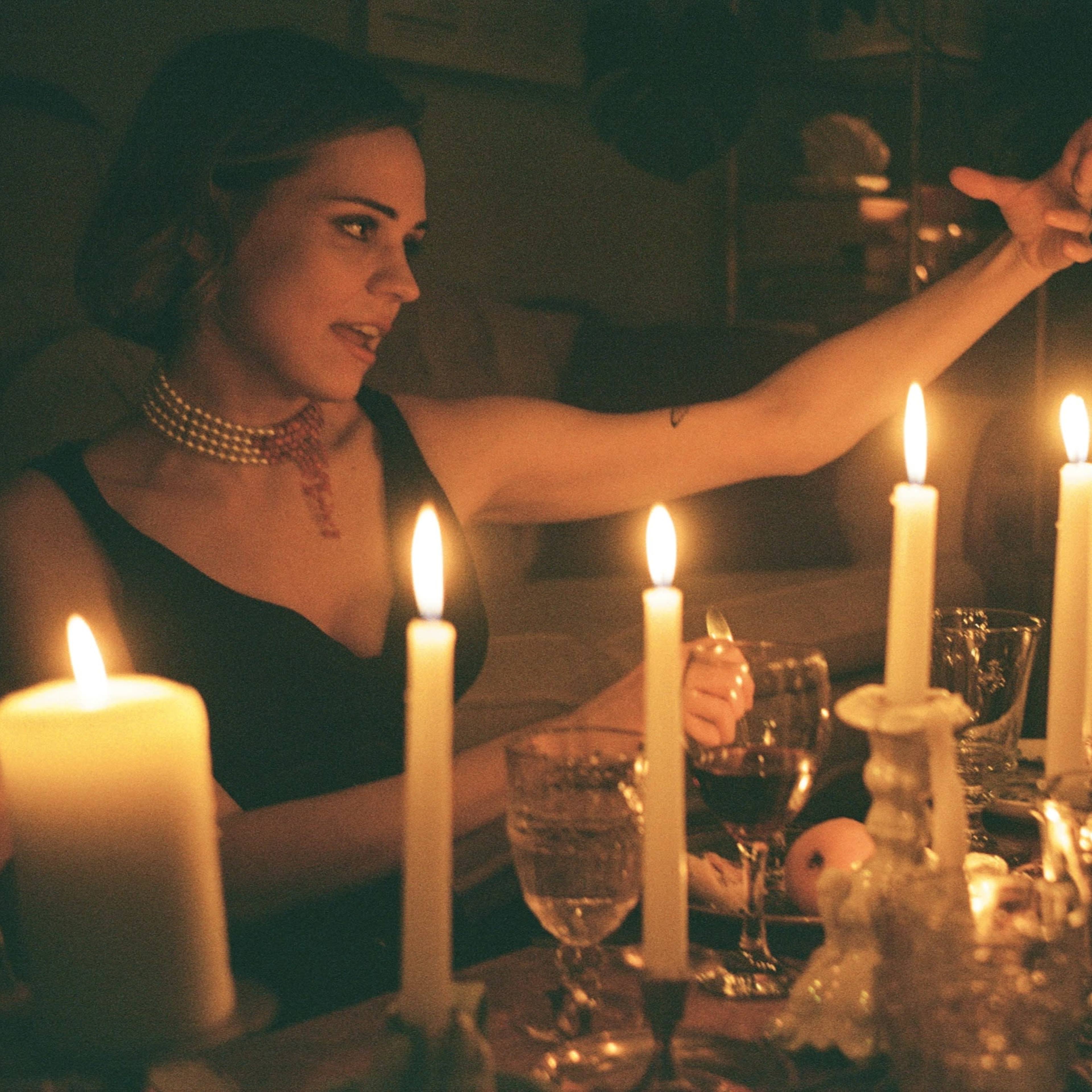 A woman gestures dramatically at a dimly lit table surrounded by candles and glassware.