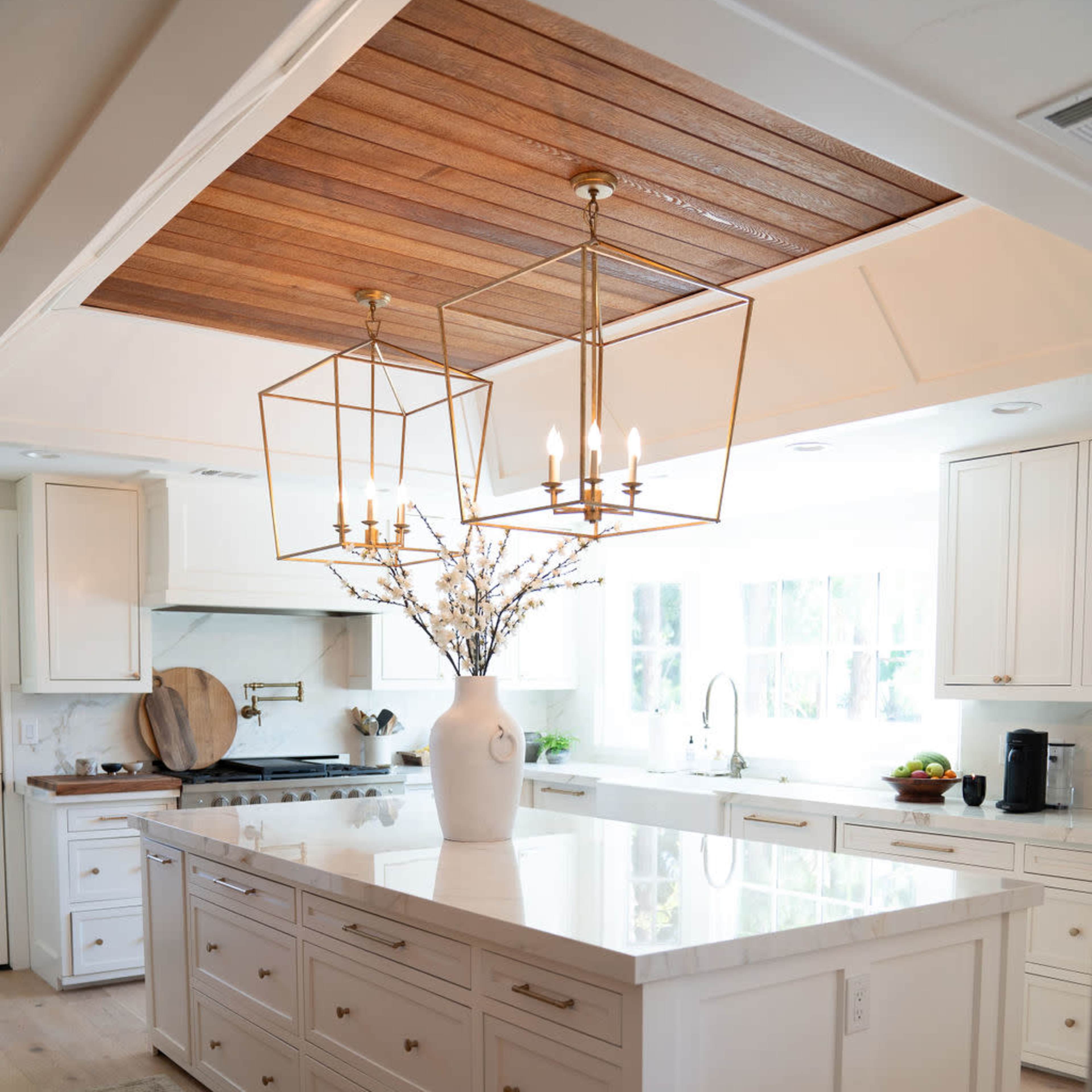 A modern kitchen features an island with multiple drawers, a large vase with branches as a centerpiece, and pendant lighting above a wooden ceiling.