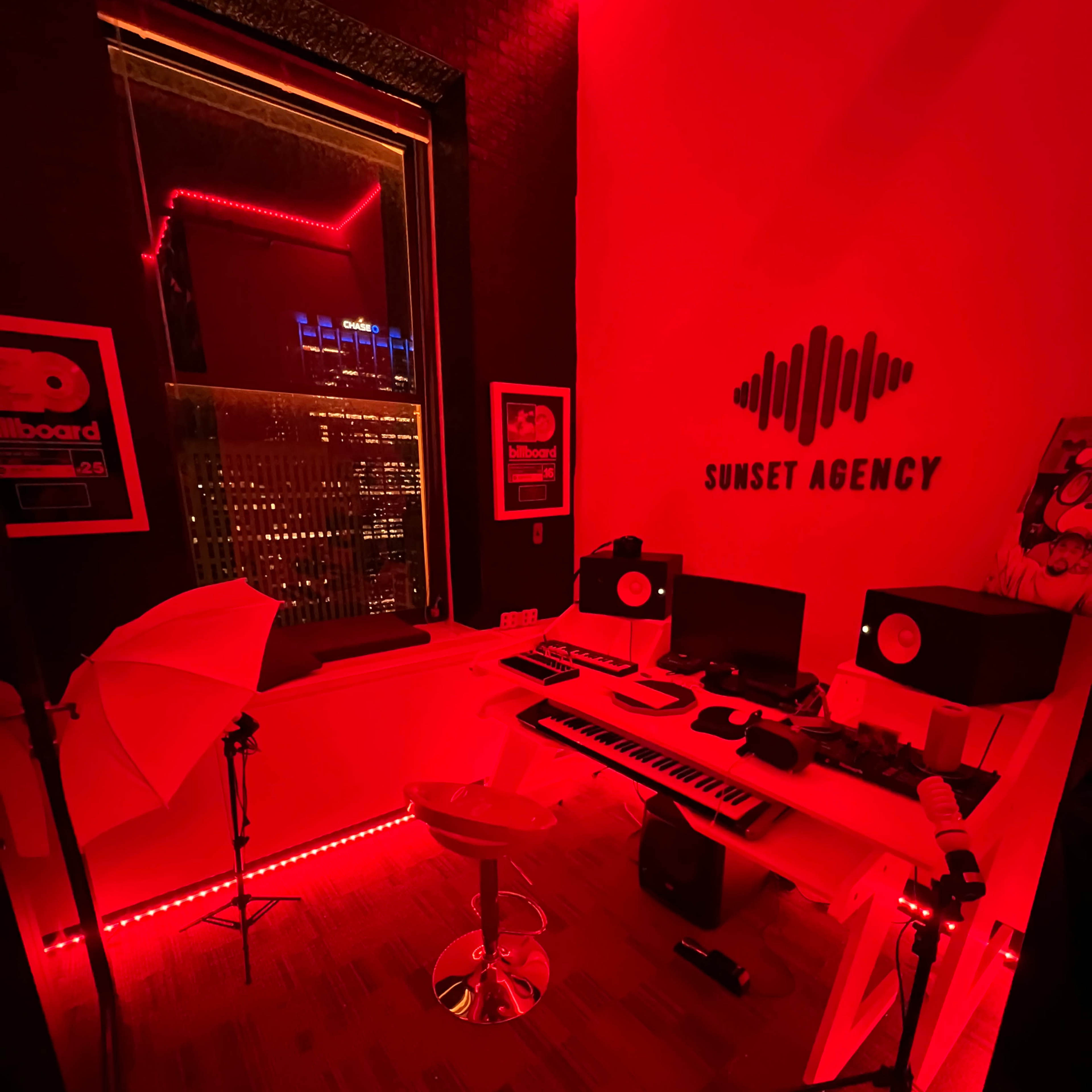 A music production studio features a white desk with a keyboard, speakers, and recording equipment, illuminated by red LED lights.