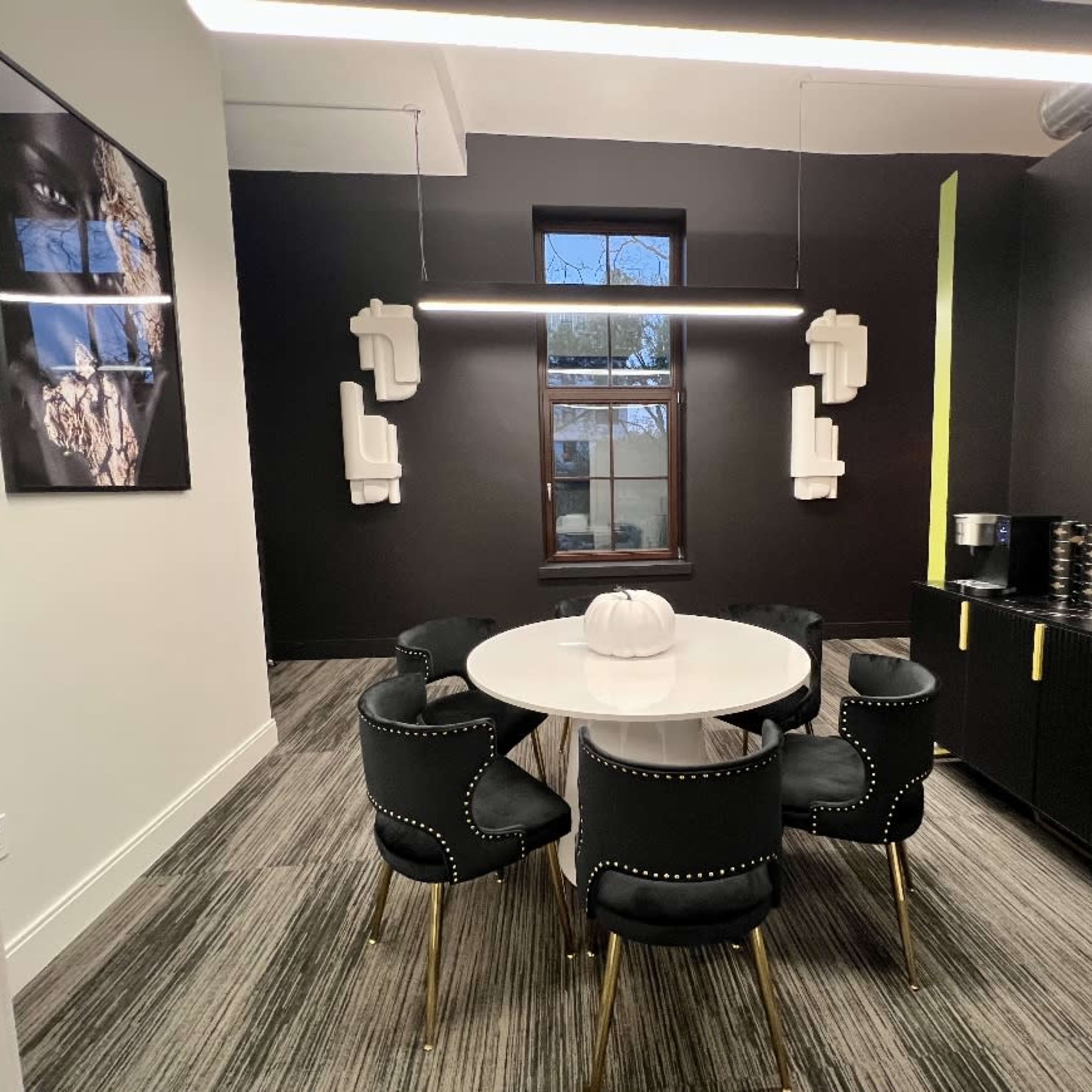 A modern meeting room features a circular white table surrounded by six black chairs with gold accents, along with a wall-mounted television and artwork.