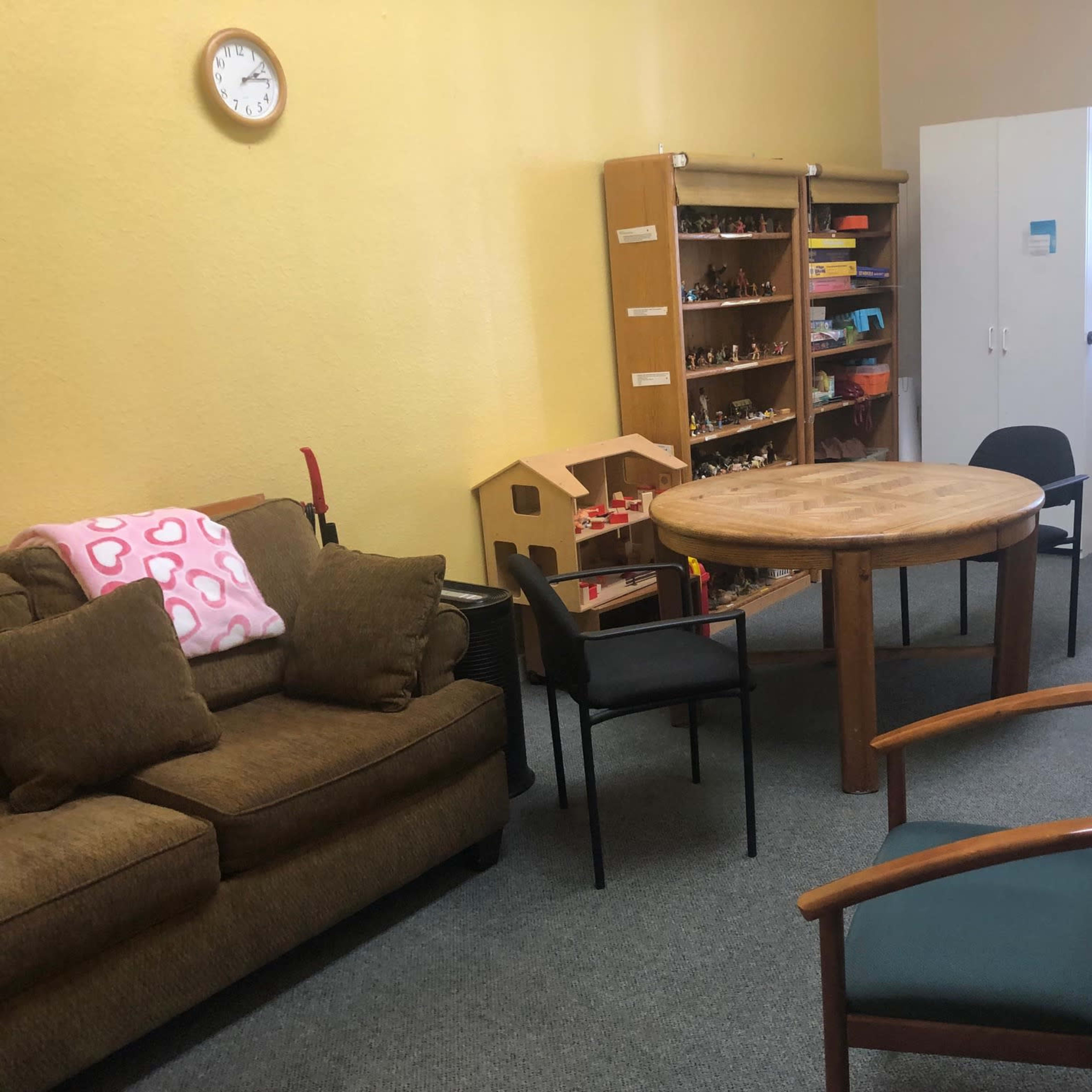 A small room with a circular table surrounded by several chairs, a couch, a bookshelf filled with toys, and a white cabinet.