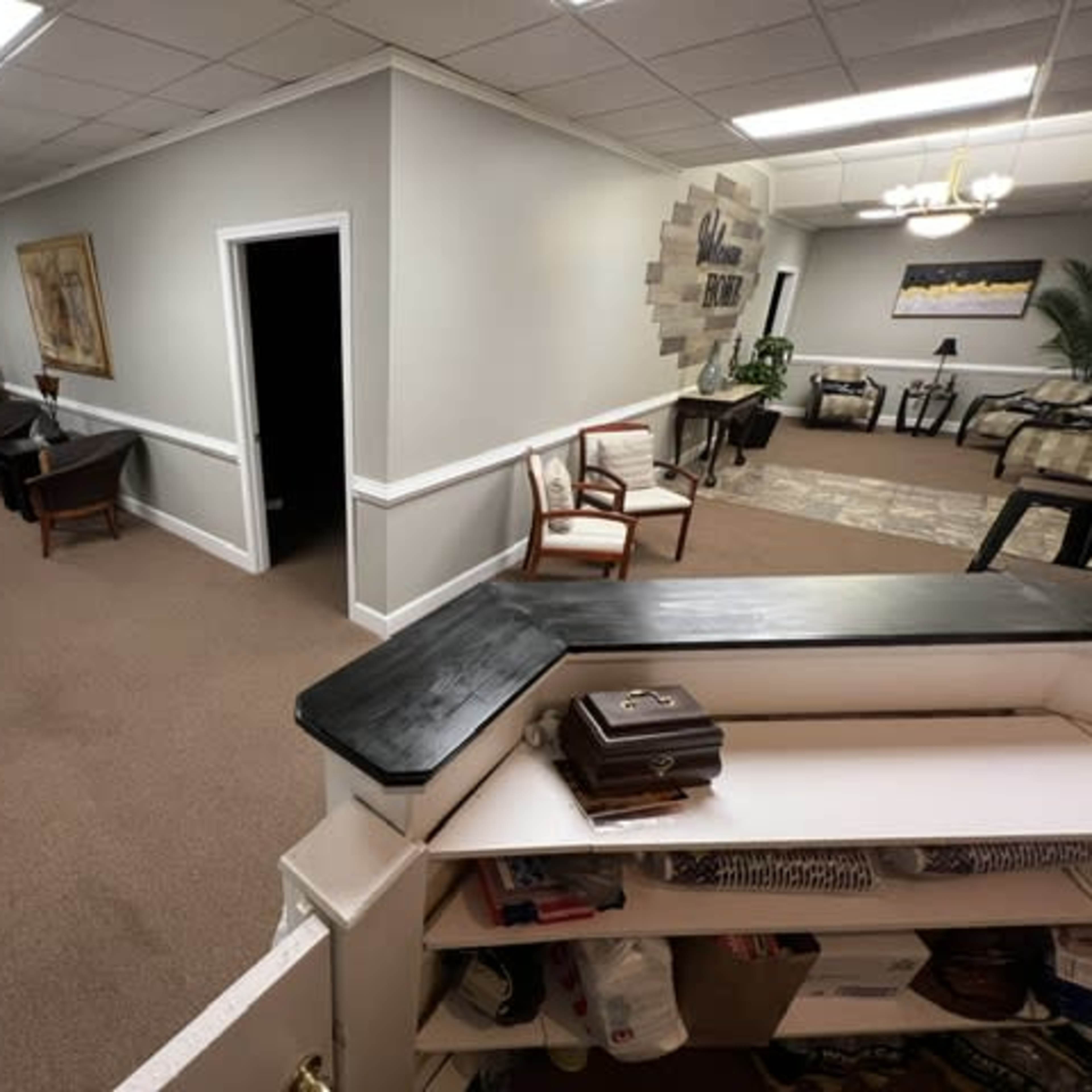 A corridor with chairs along the walls, leading to various rooms and a reception area with a desk.