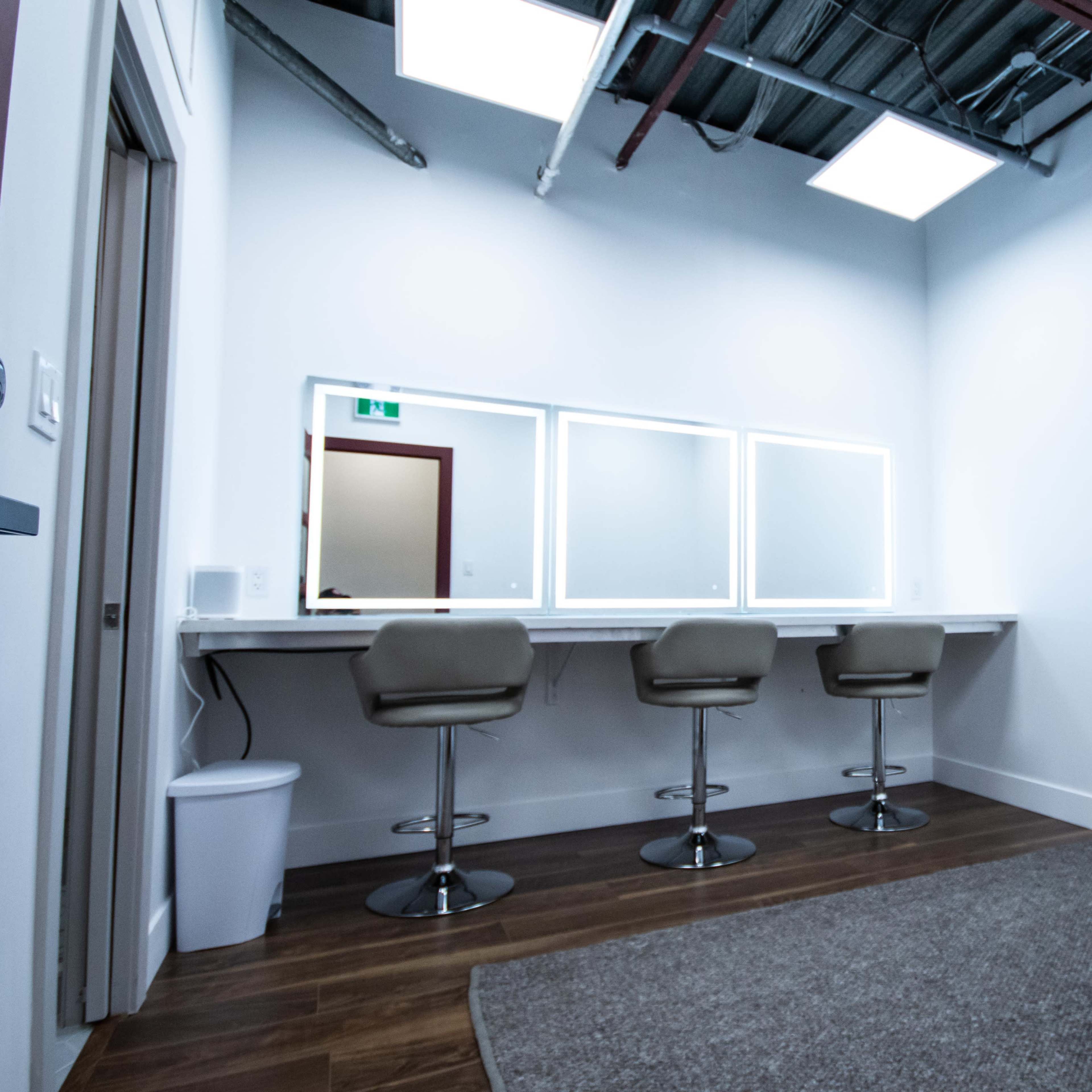 A minimalist makeup room featuring three bar stools at a long counter with three illuminated mirrors, a wastebasket, and a carpeted floor.