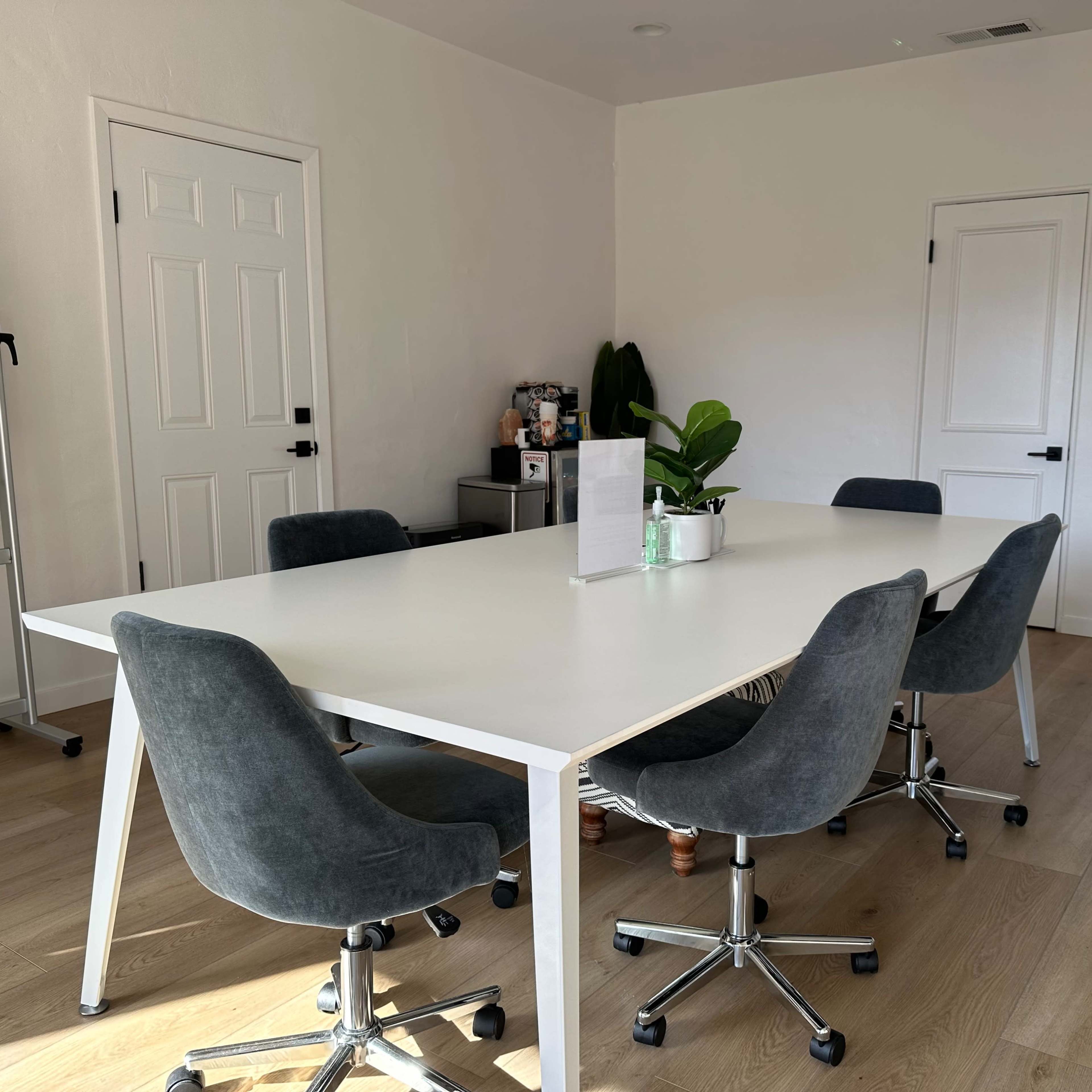 A modern conference room features a large white table with four gray chairs and a small plant on the table, set in a bright, minimalistic space.
