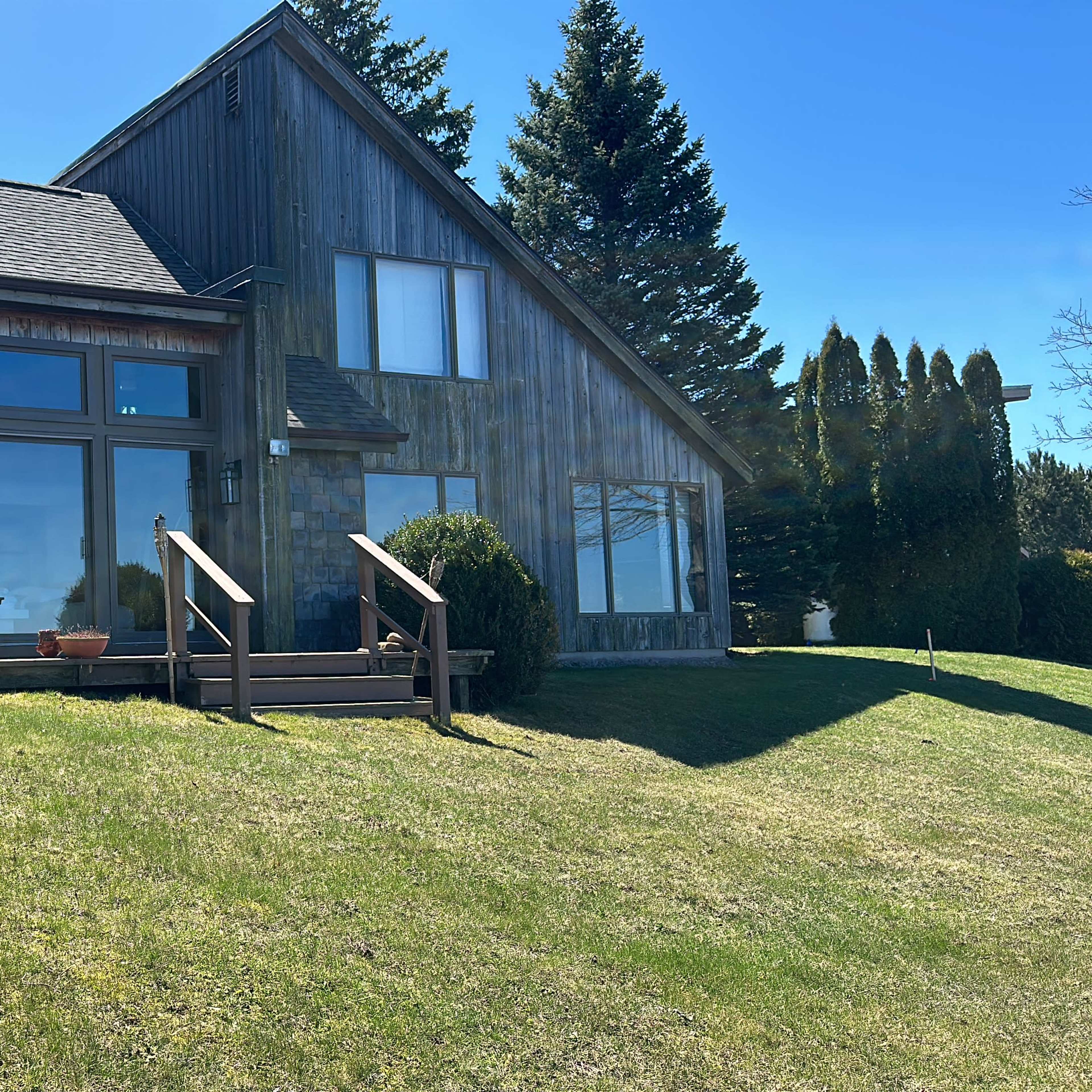 A large wooden house with large windows and a deck, set on a grassy lawn under a clear blue sky.