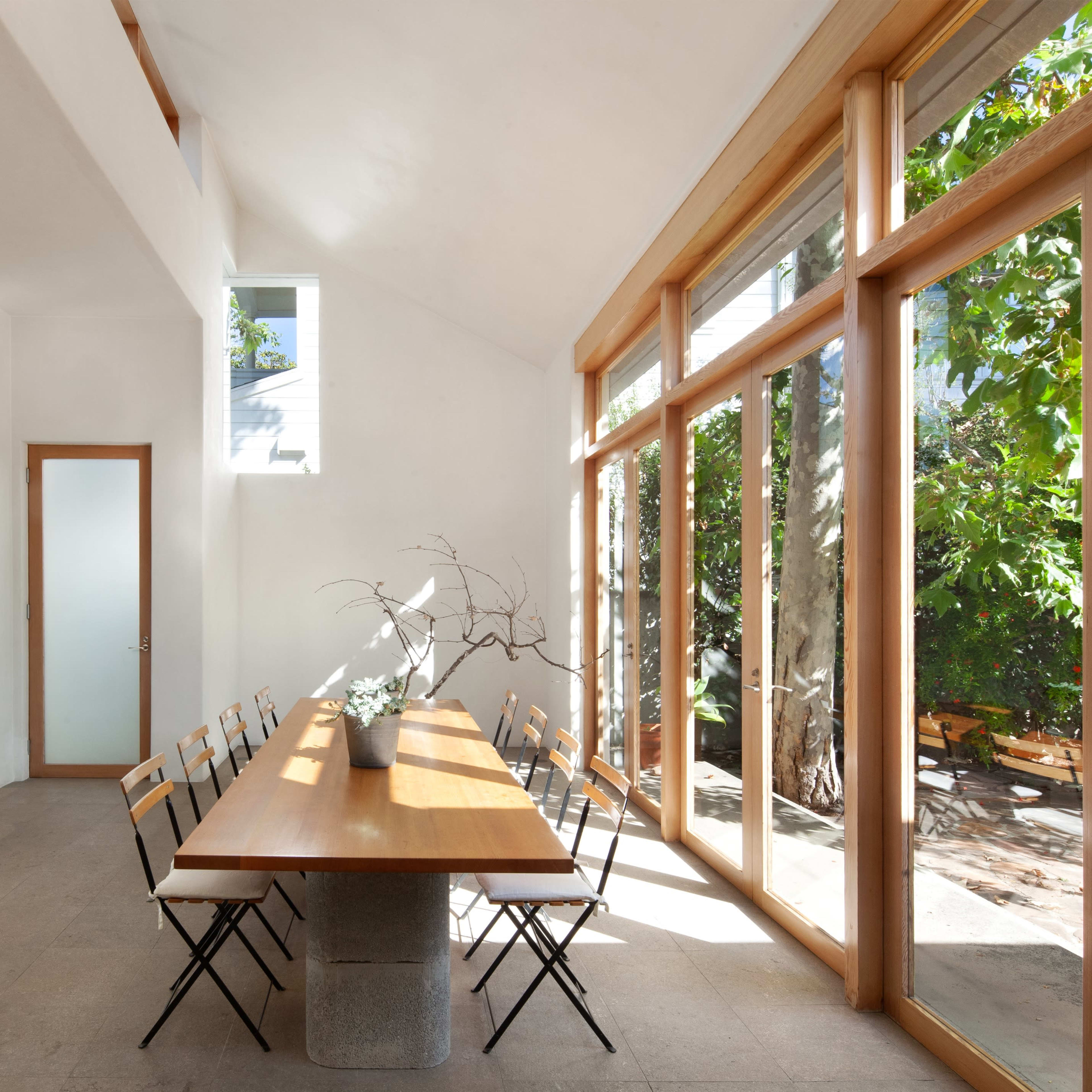 A minimalist dining area with a long wooden table surrounded by black chairs, large windows letting in natural light, and a view of greenery outside.