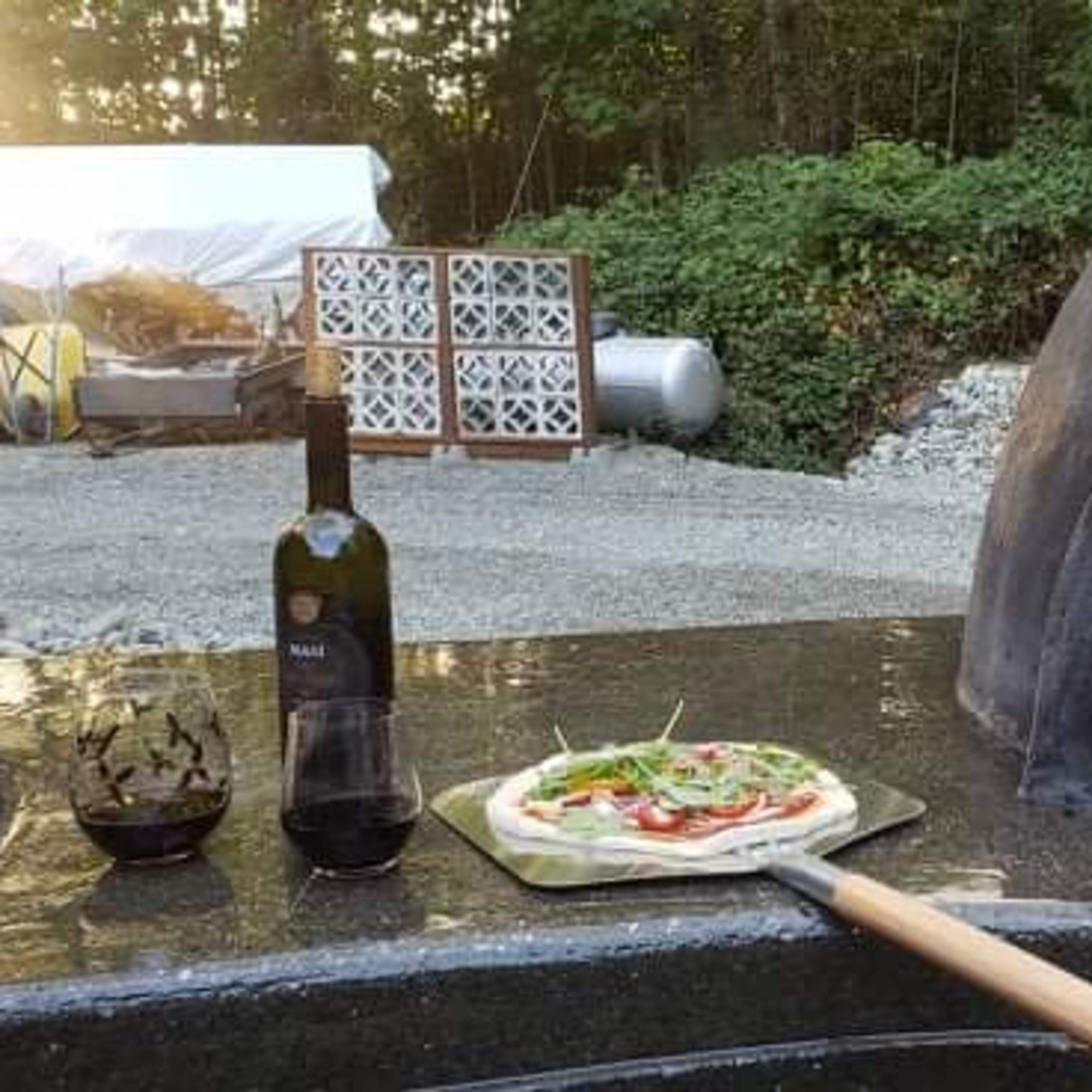 A pizza is being placed into a wood-fired oven with a bottle of olive oil and a glass of wine nearby.