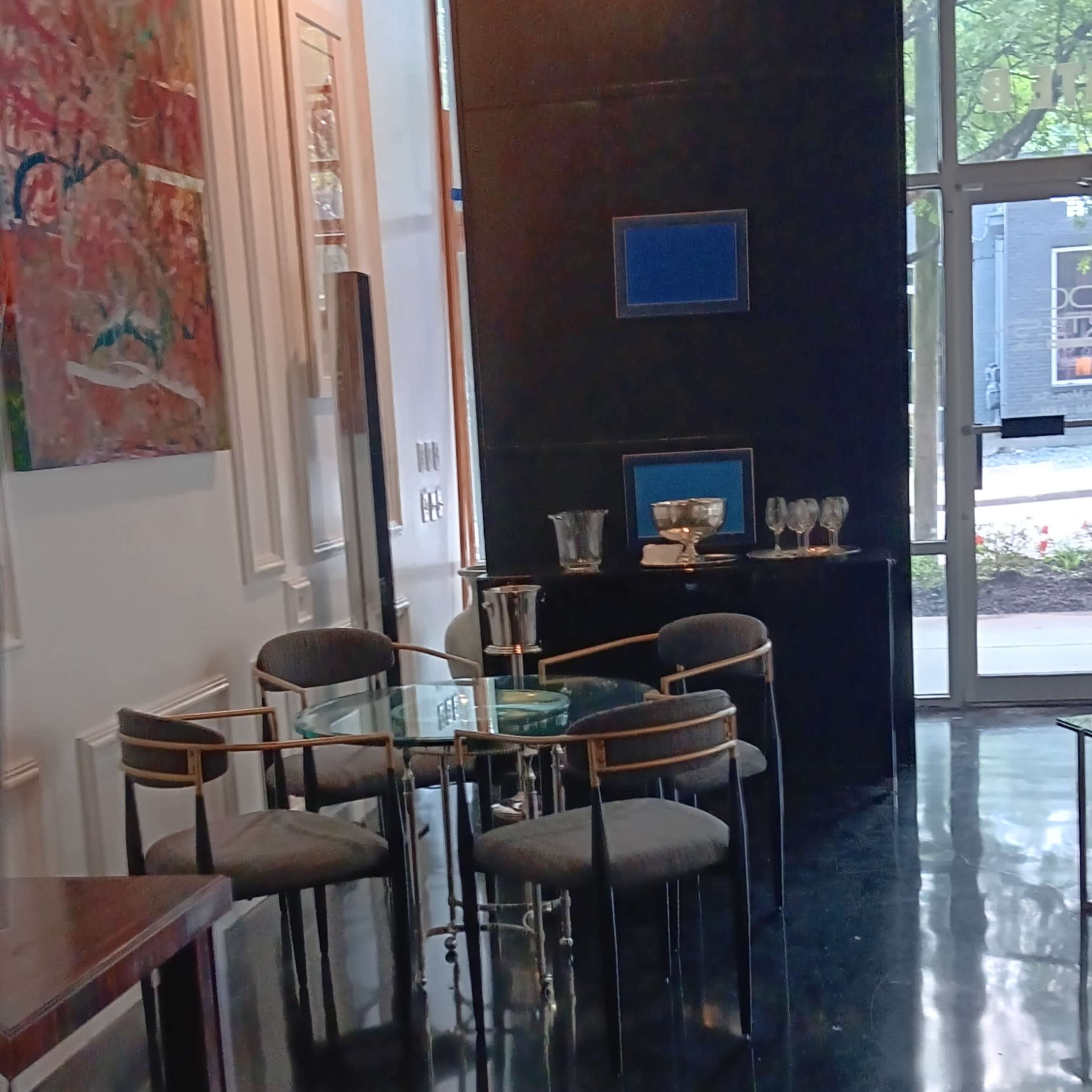 A modern interior space with a glass dining table surrounded by four upholstered chairs, artwork on the walls, and a large window facing a courtyard.