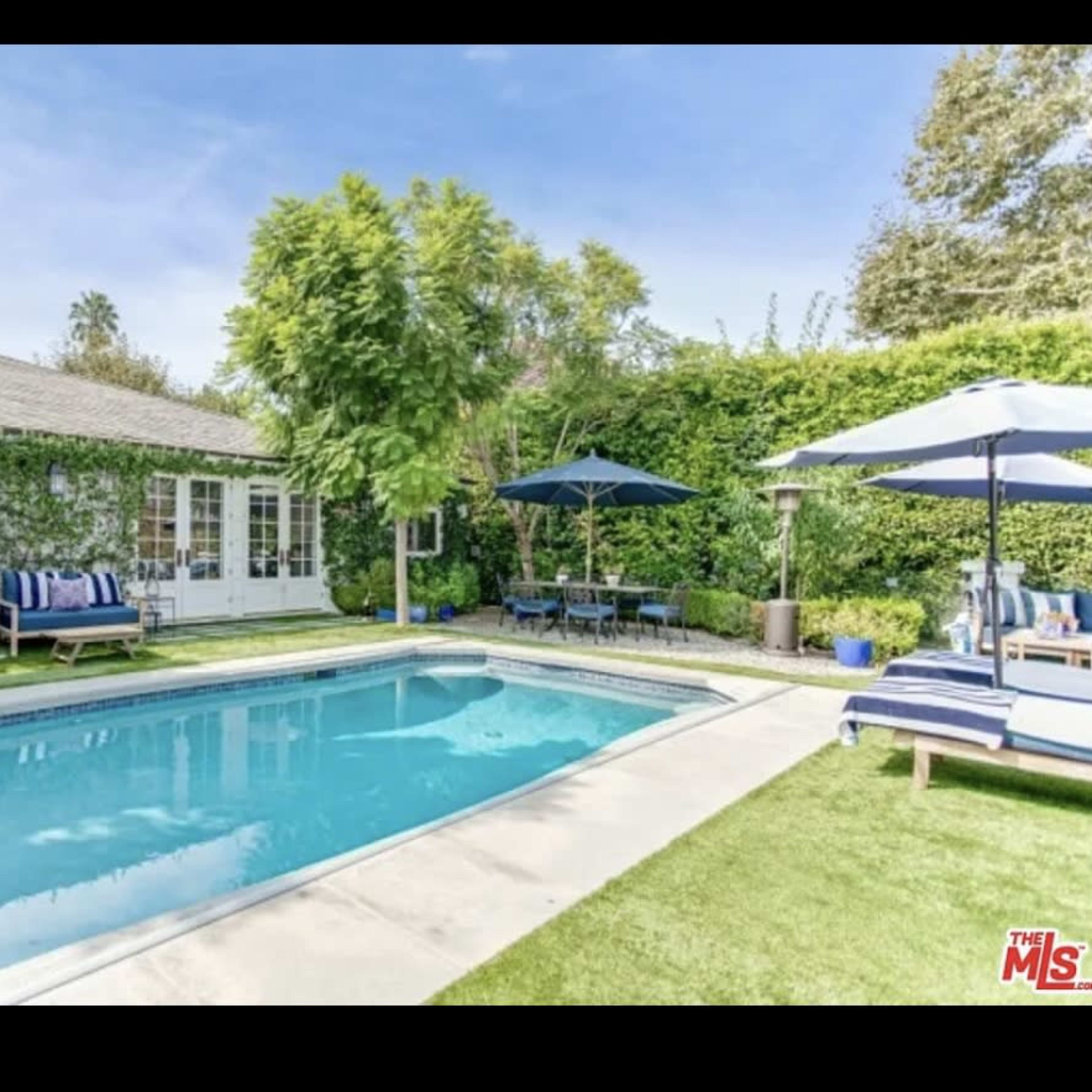 A rectangular swimming pool is surrounded by a landscaped yard featuring greenery, lounge chairs, and tables with umbrellas.