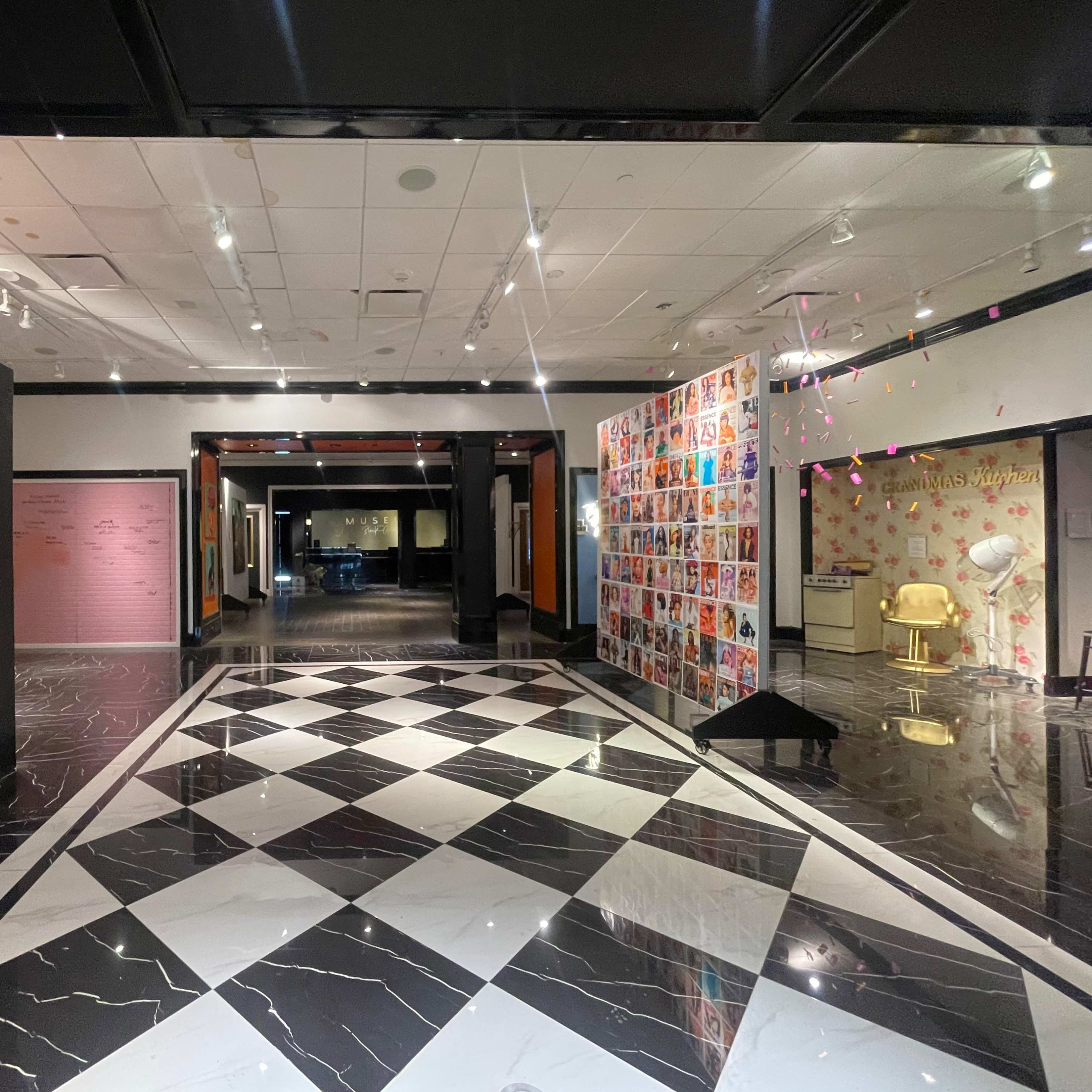A spacious gallery with a checkered black and white floor, featuring walls adorned with colorful photographs and text.