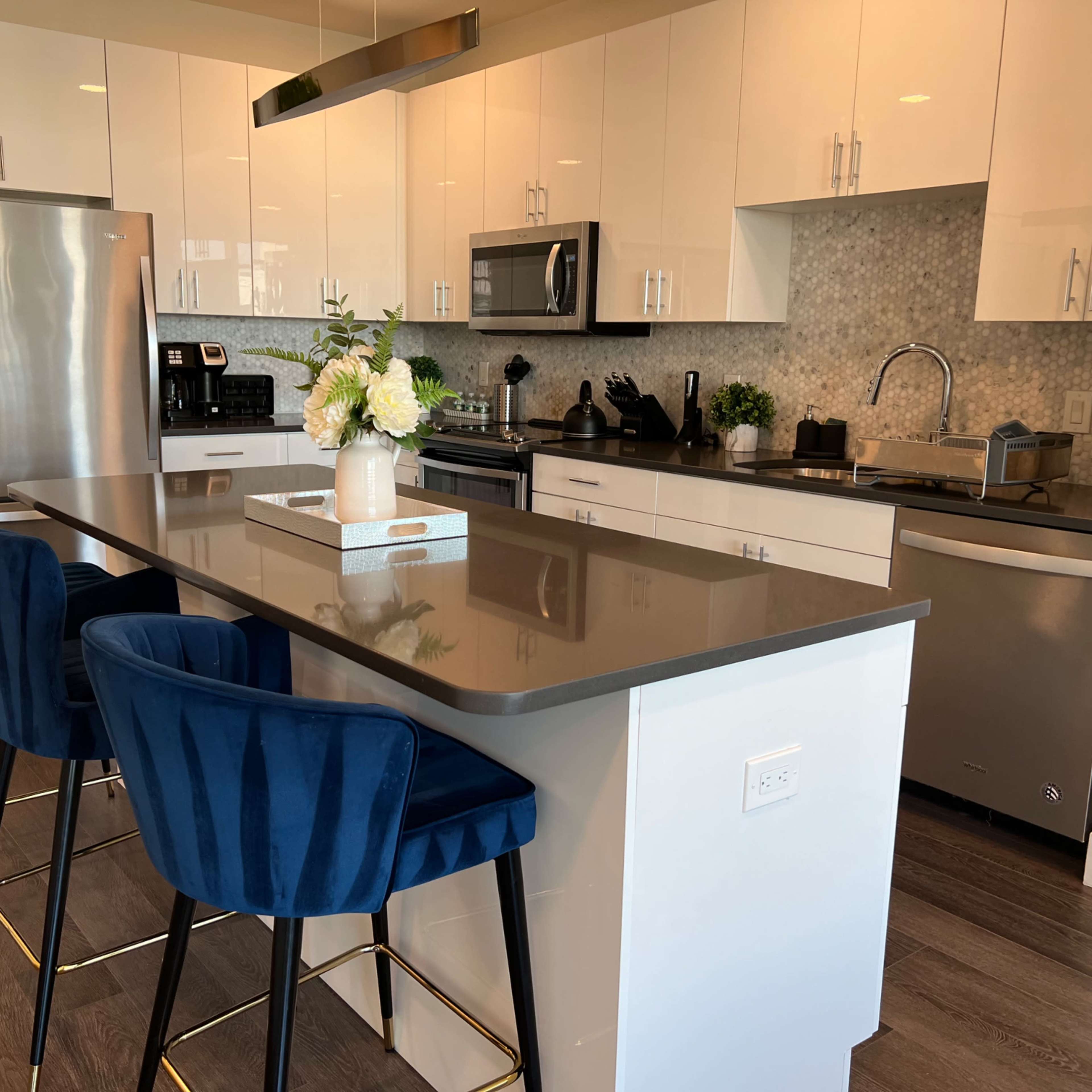A modern kitchen with a large island featuring four blue stools, stainless steel appliances, and white cabinetry.